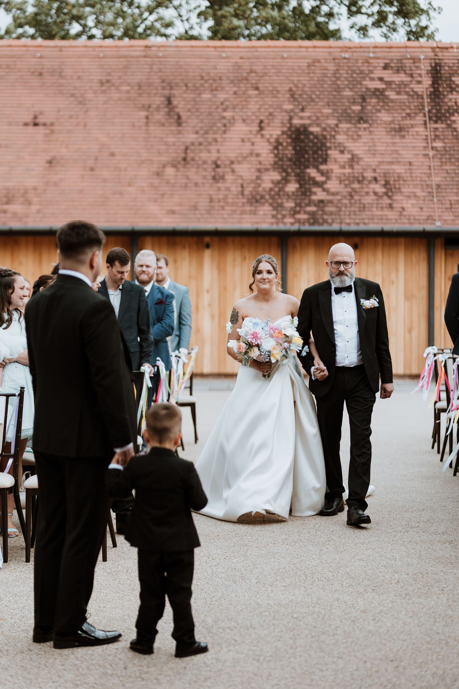 Bride walks down the aisle with her father at an outdoor barn wedding ceremony, holding a pink bouquet.