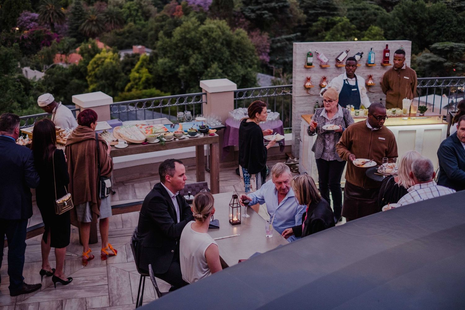 Elegant outdoor rooftop cocktail party at dusk with guests dining and mingling near a decorative display board.