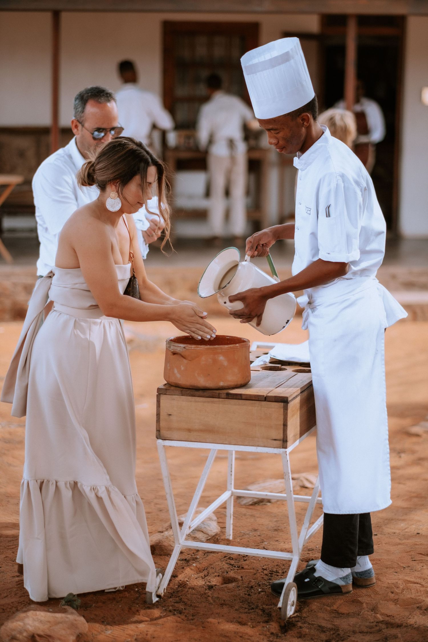 Chef serving food from a wooden cart to a woman in a beige dress at an outdoor dining event.