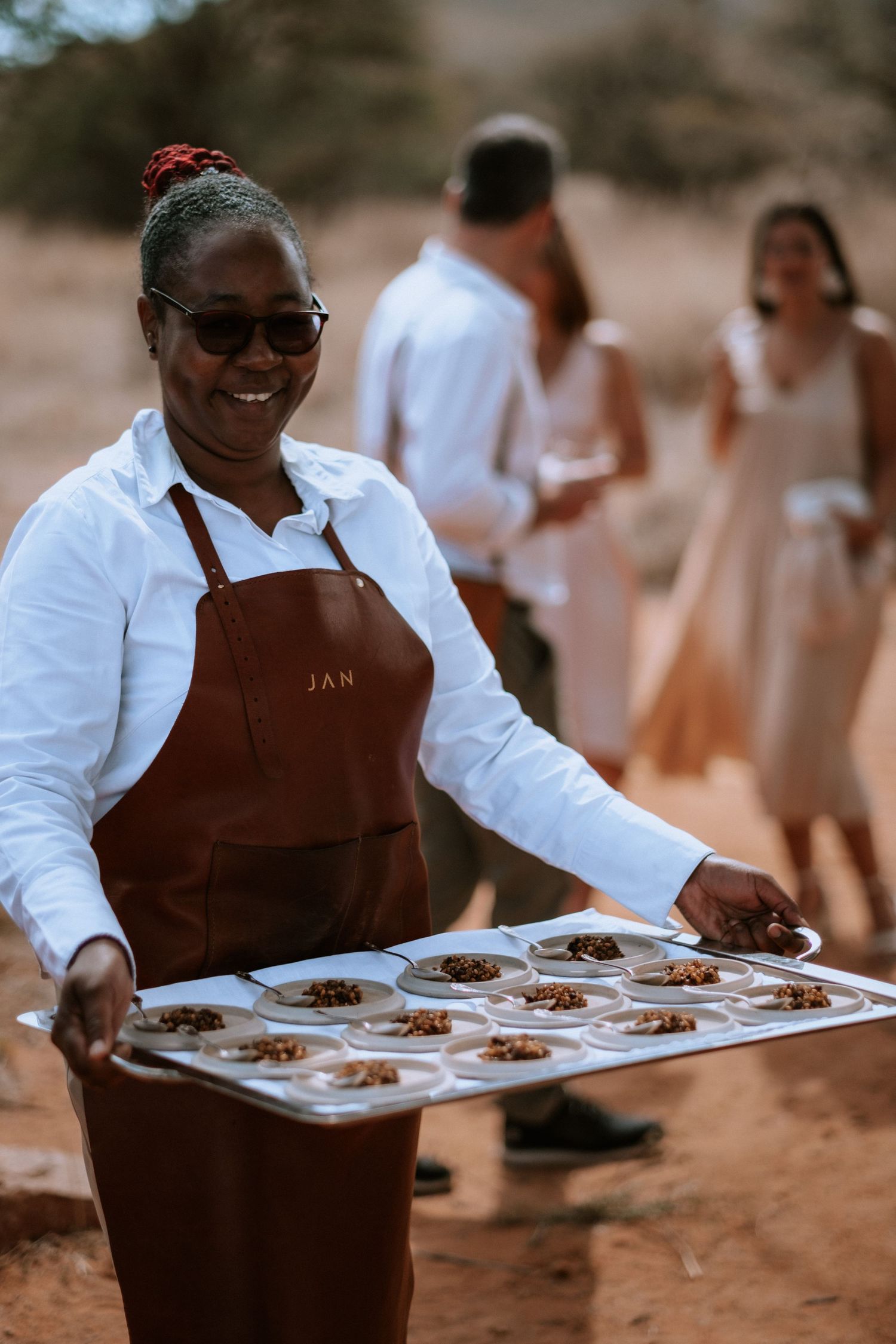Smiling waitress in brown JAN apron holds tray of appetizer plates outdoors at a desert event.