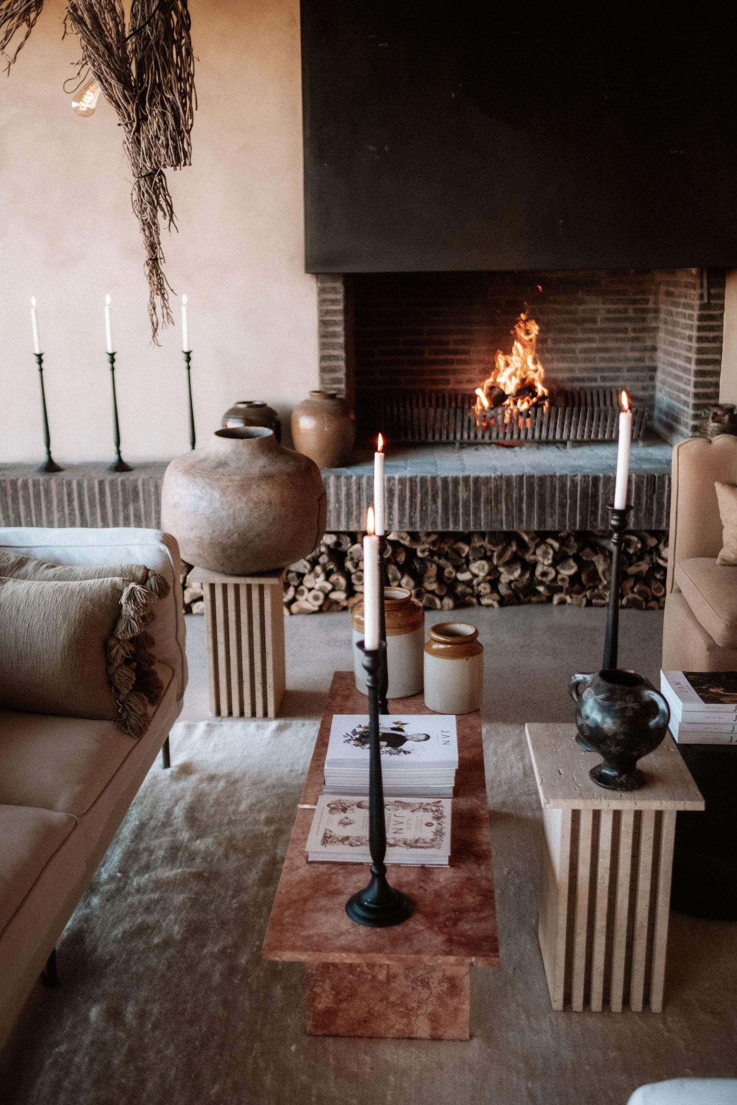 Cozy rustic living room with lit fireplace, taper candles, earthy decor, stacked firewood, and neutral tones.