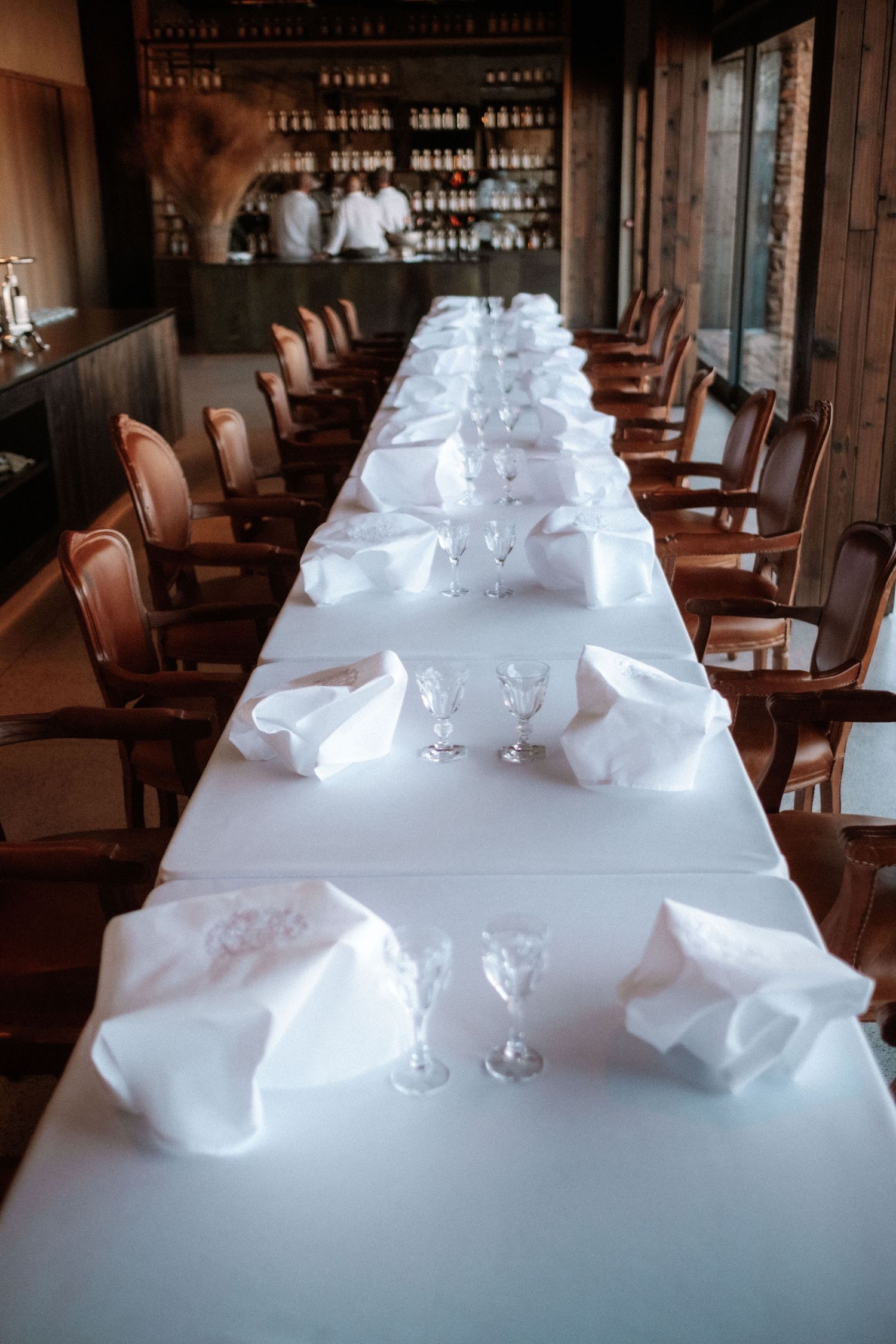Elegant long dining table set with white linens, crystal glasses, and folded napkins in a rustic restaurant setting.