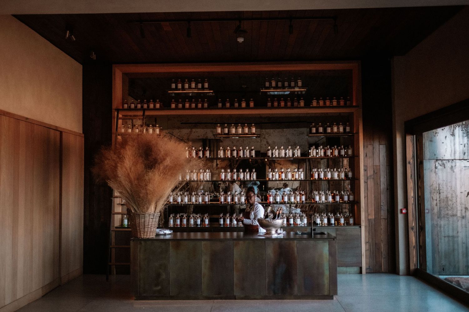 Rustic bar with shelves of bottles, a bartender, and pampas grass decor in a warm wooden interior.