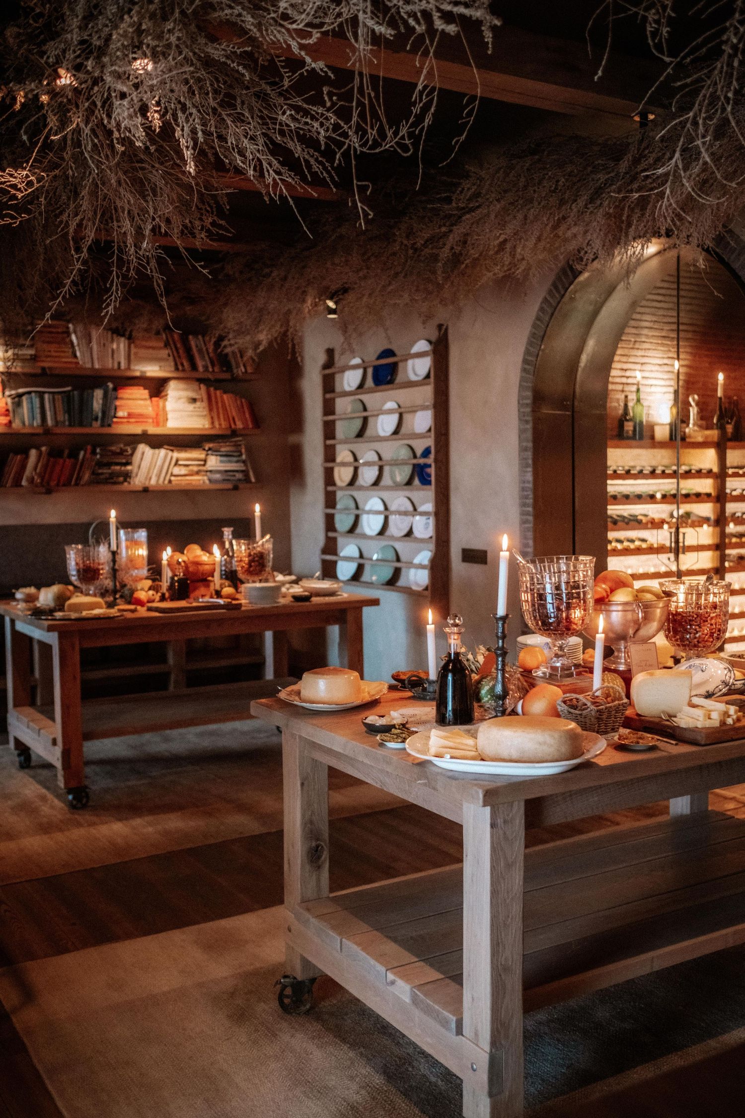 Rustic kitchen dining area with candlelit wooden tables, displayed plates, dried botanicals, and warm ambient lighting.