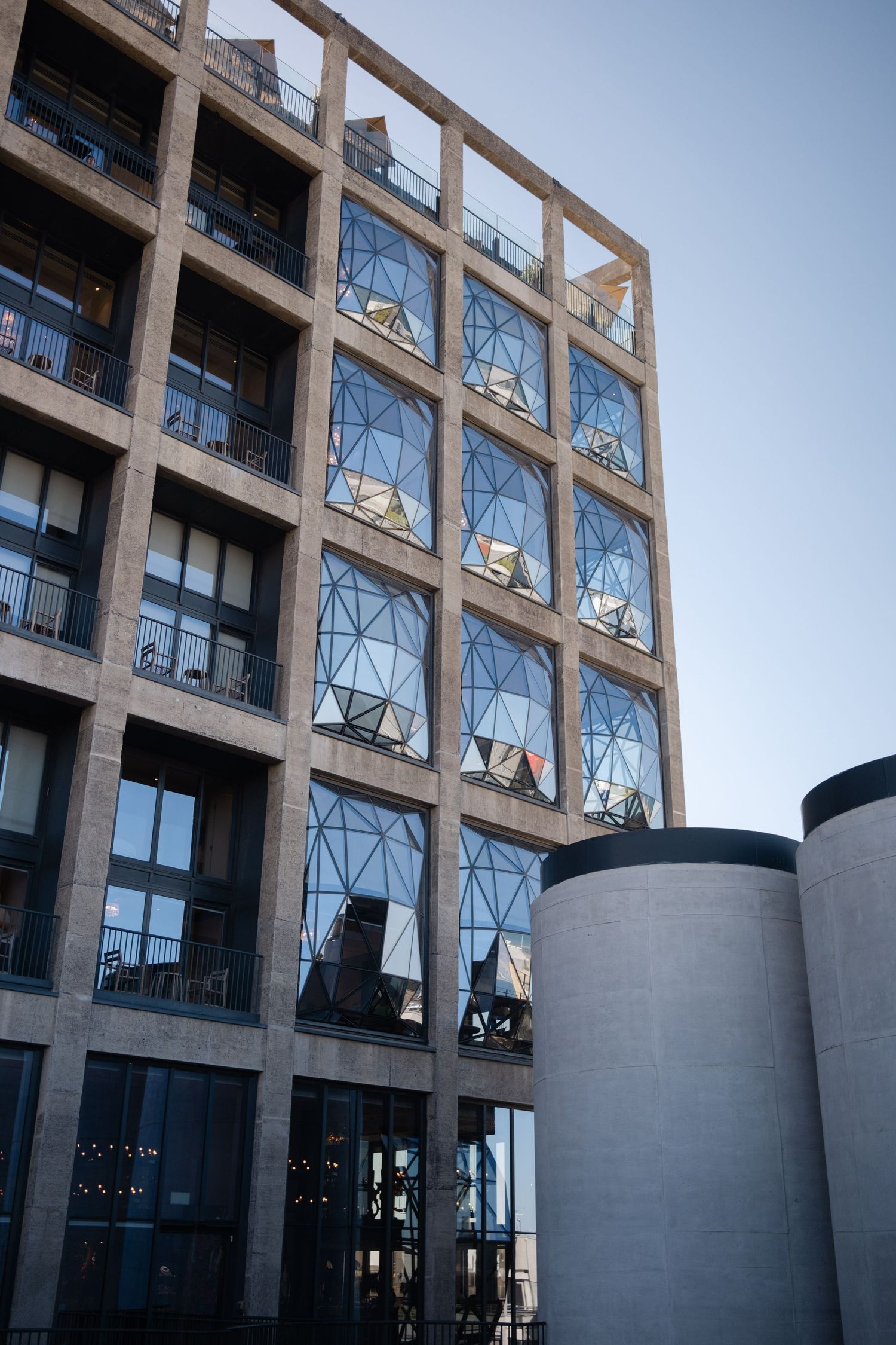 Modern multi-story building with geometric glass windows and cylindrical concrete columns against a clear blue sky.