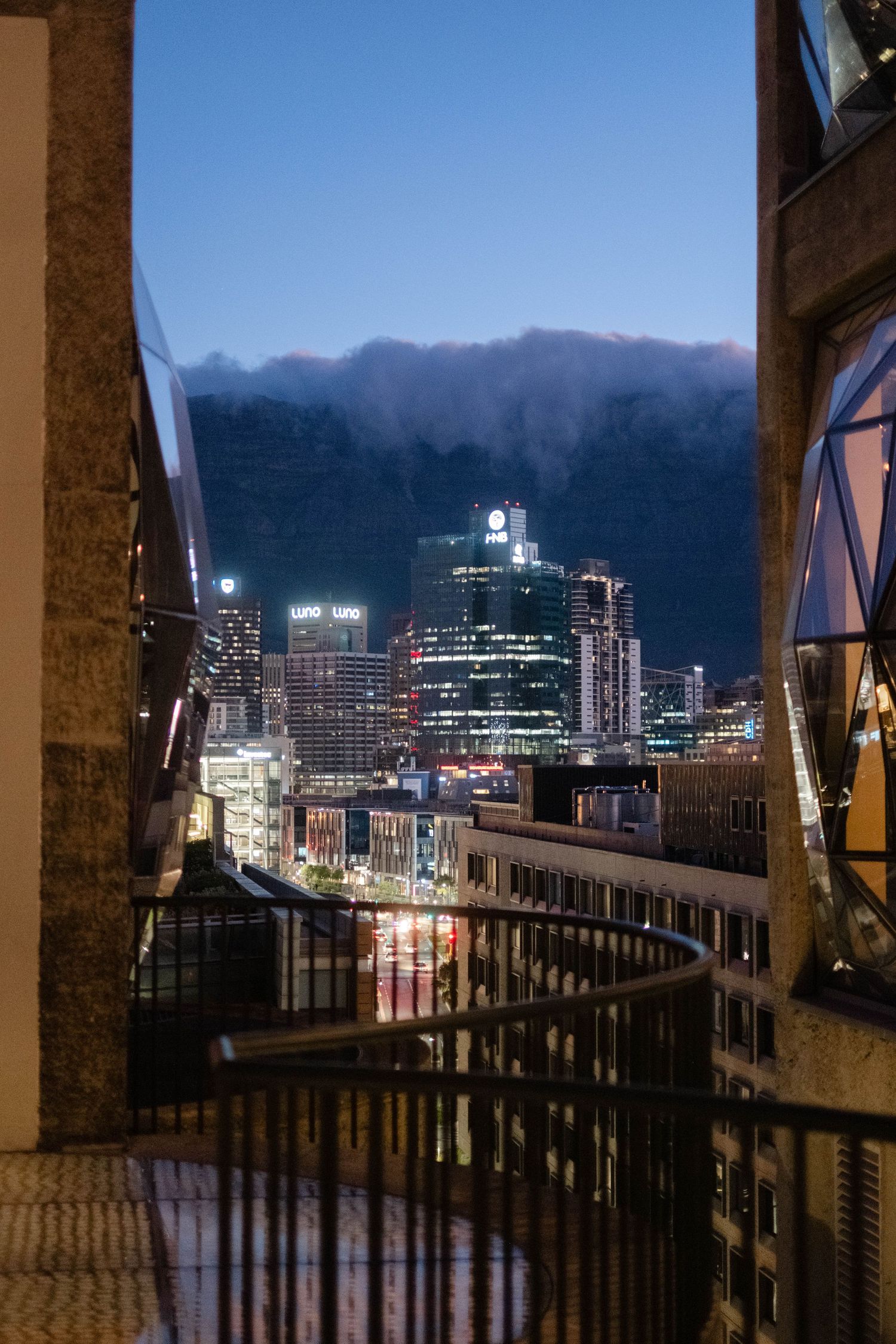 Night cityscape viewed from a balcony with illuminated skyscrapers and a mountain backdrop under a blue dusk sky.