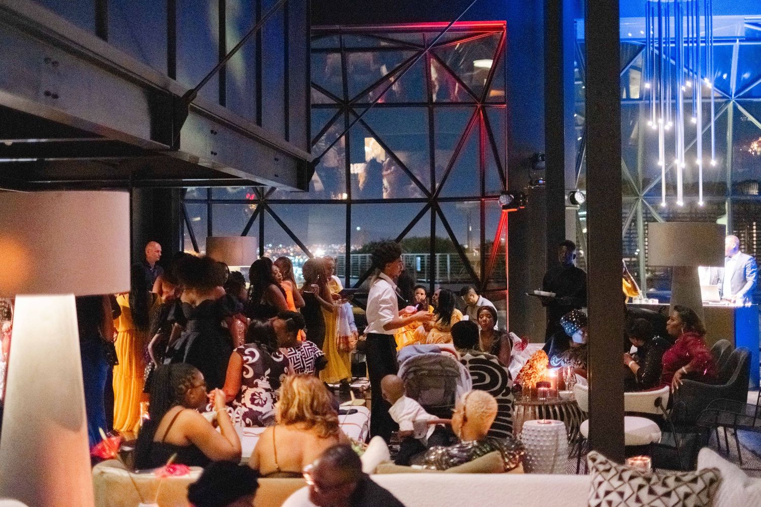 Lively rooftop bar with guests socializing under industrial steel beams and large windows at night.