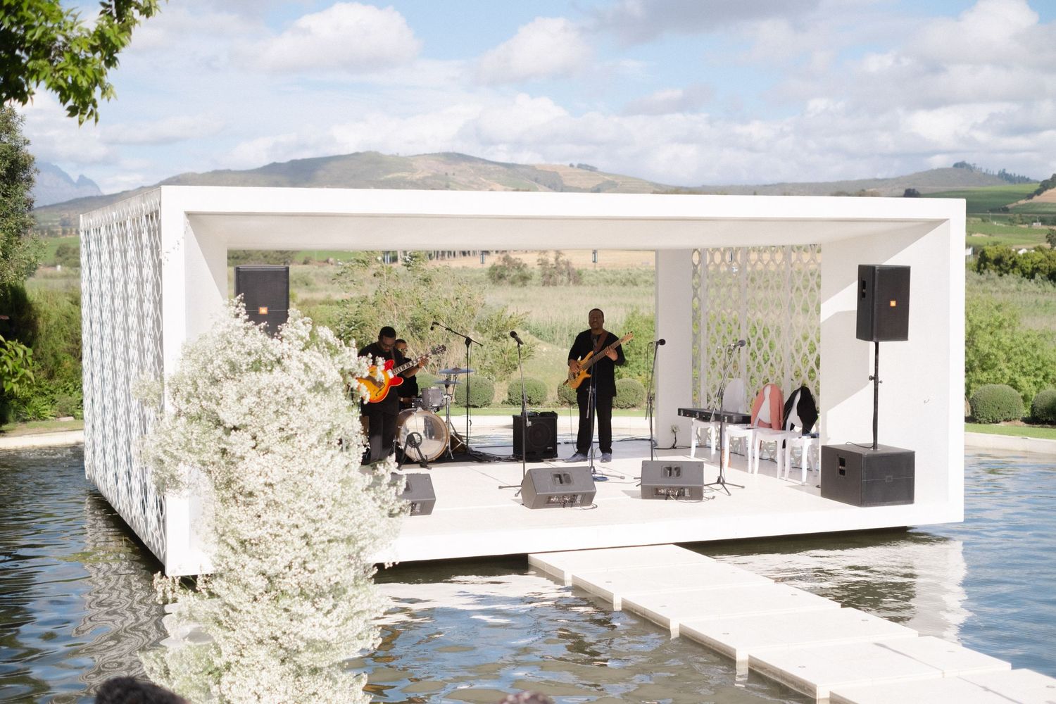 Live band performing on a floating white stage over a calm lake with scenic hills in the background.