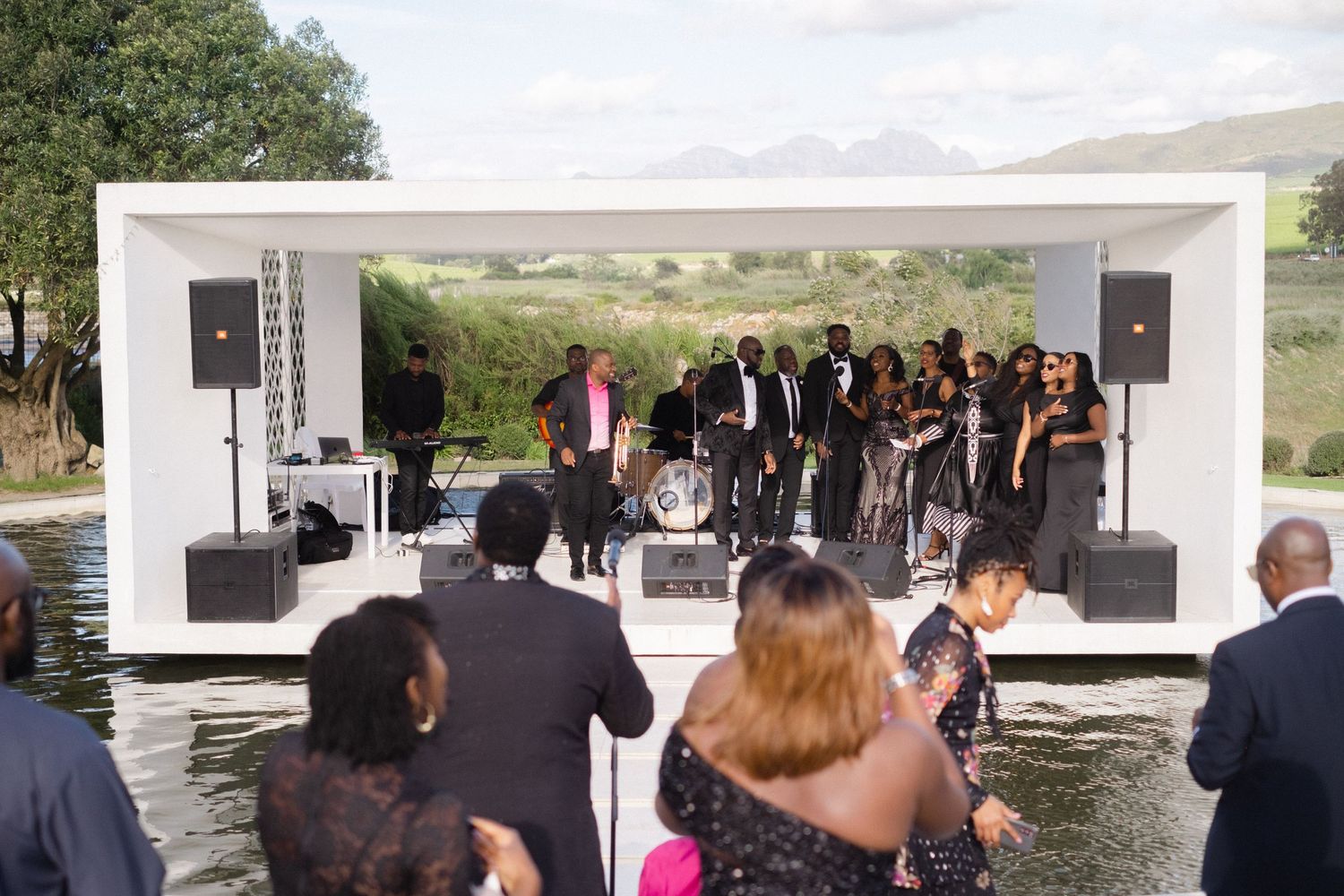 Live band performing on a floating stage over water at an outdoor event with mountains in the background.