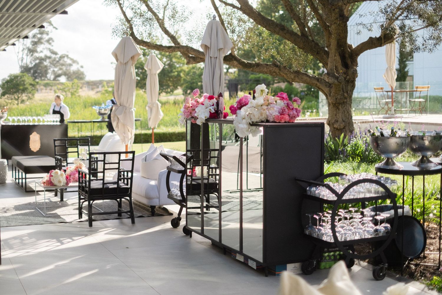 Elegant outdoor wedding venue with black chairs, pink floral arrangements, and white umbrellas on a sunny patio.