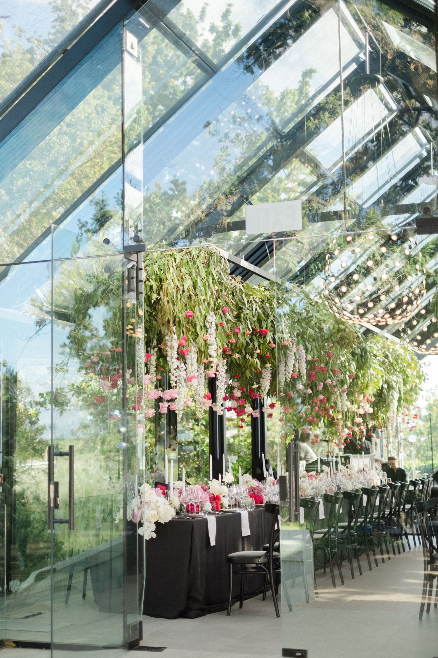 Elegant greenhouse wedding venue with floral ceiling installations, dark draped tables, and glass walls overlooking lush greenery.