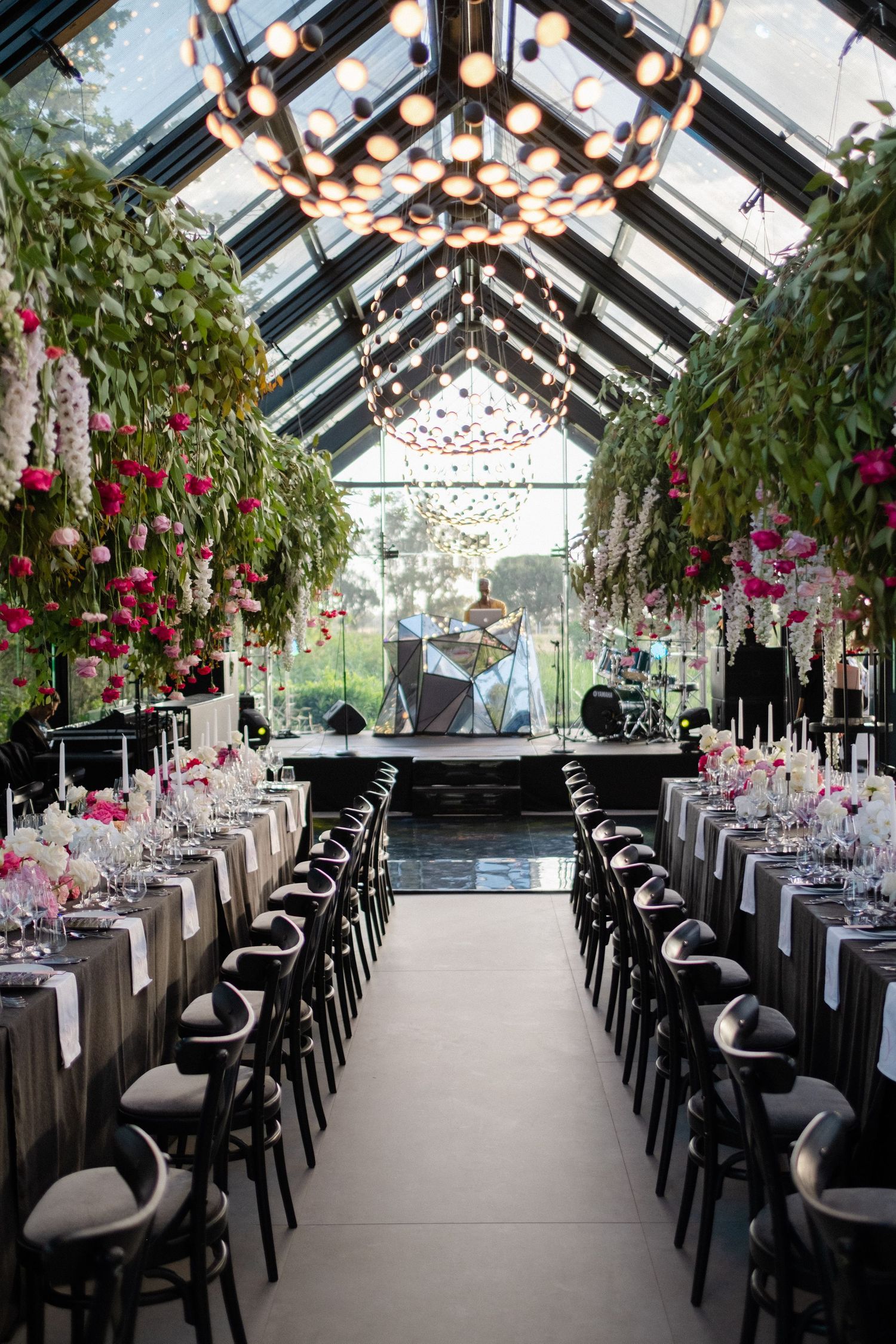 Elegant greenhouse wedding reception with hanging florals, long tables, black chairs, and chandelier lighting.