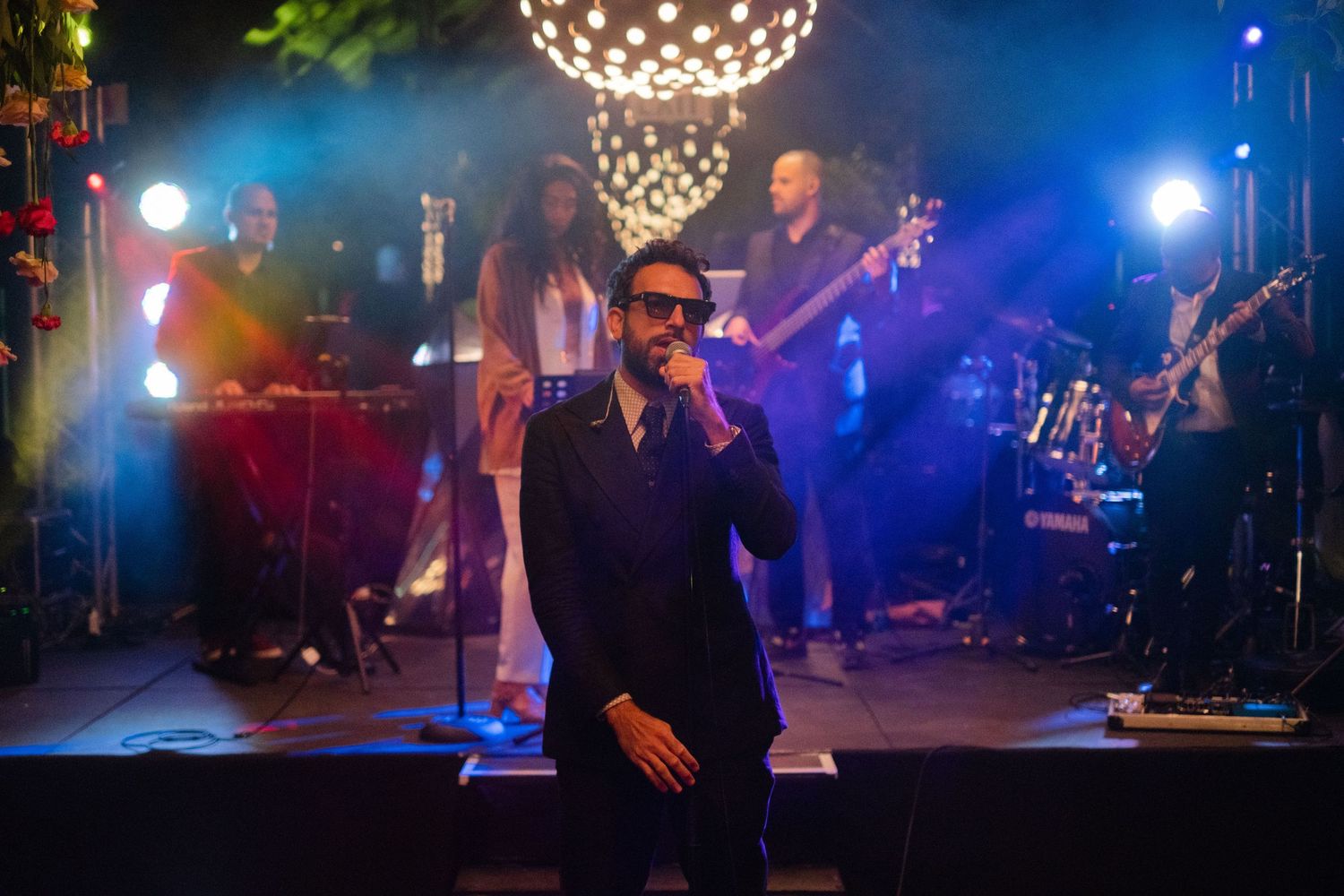 A male singer performs on stage with a live band under colorful lights at an evening concert event.