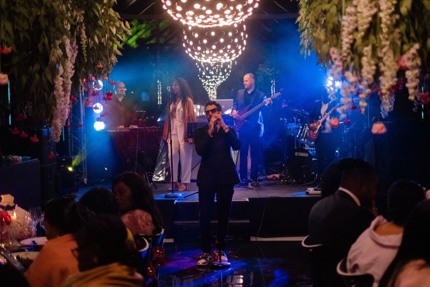 Live band performing on a decorated stage with lush greenery, warm lights, and an engaged audience at an indoor event.