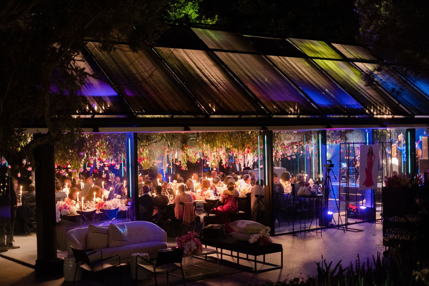 Elegant greenhouse dining event at night with colorful lighting and guests seated at candlelit tables.