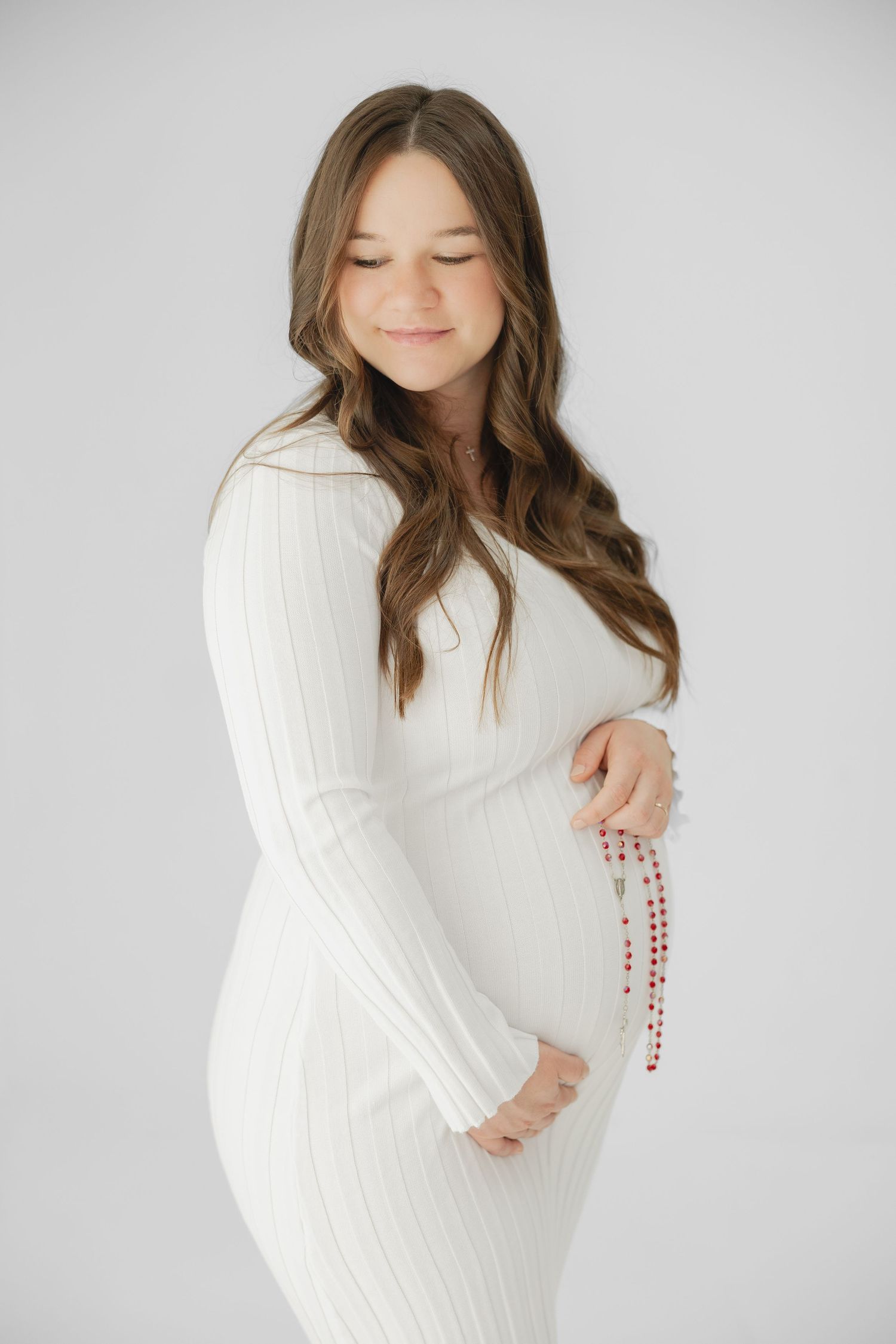 Pregnant woman in white ribbed dress holding red rosary beads against her baby bump on white background.
