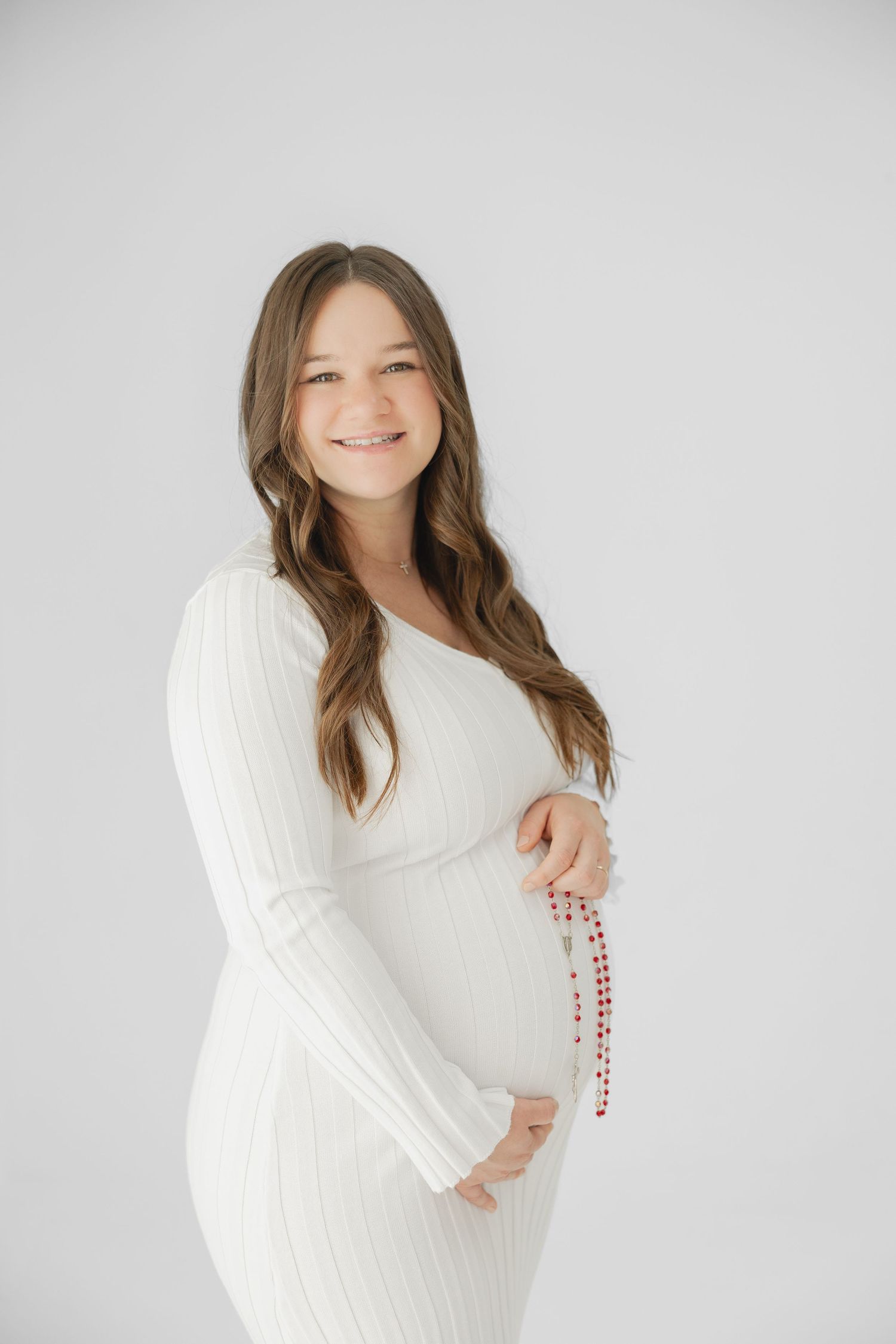 Smiling pregnant woman in white ribbed dress holding red beads over baby bump in white studio.