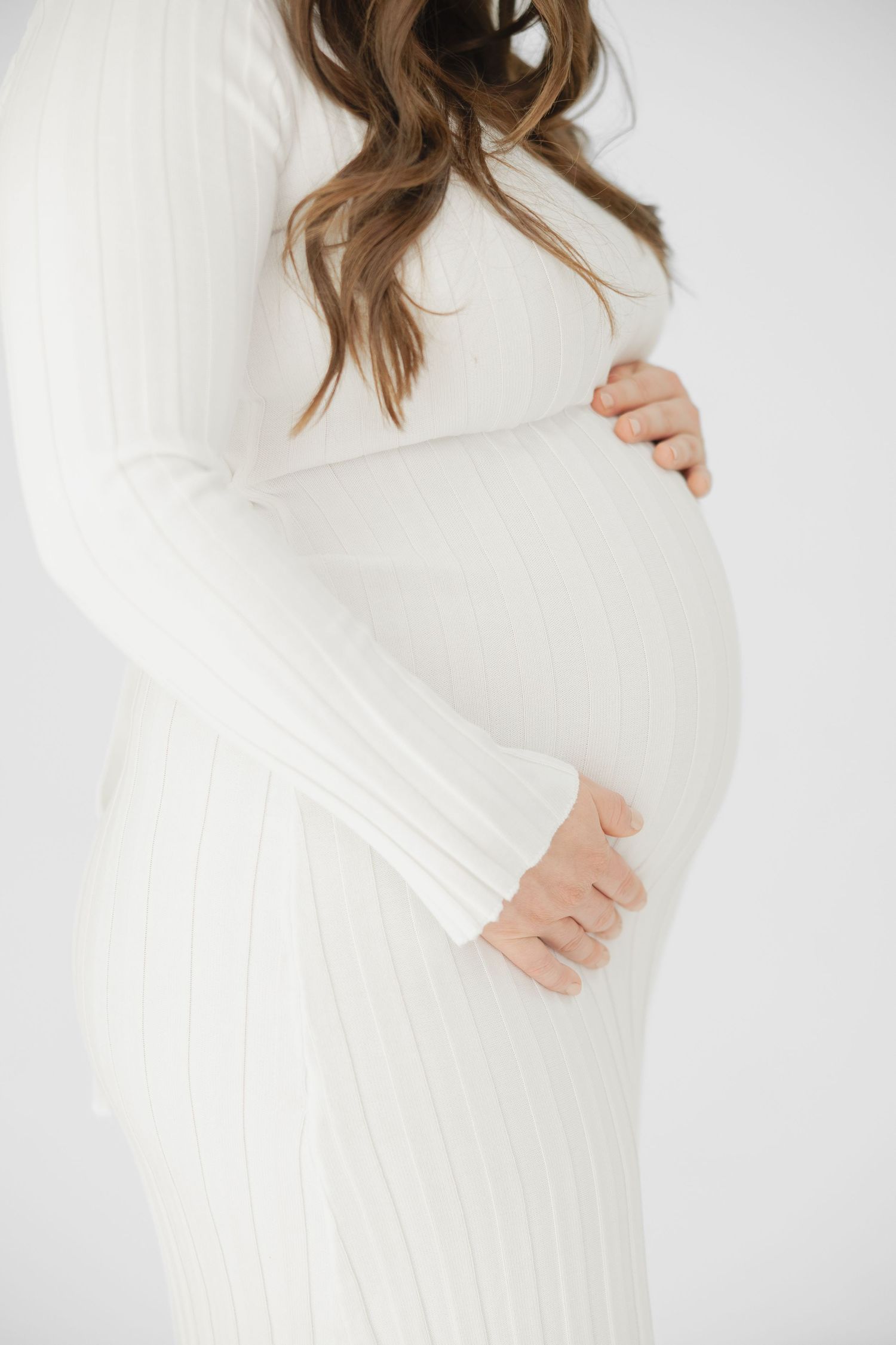 Pregnant woman in white ribbed knit dress cradling baby bump with both hands against white background.