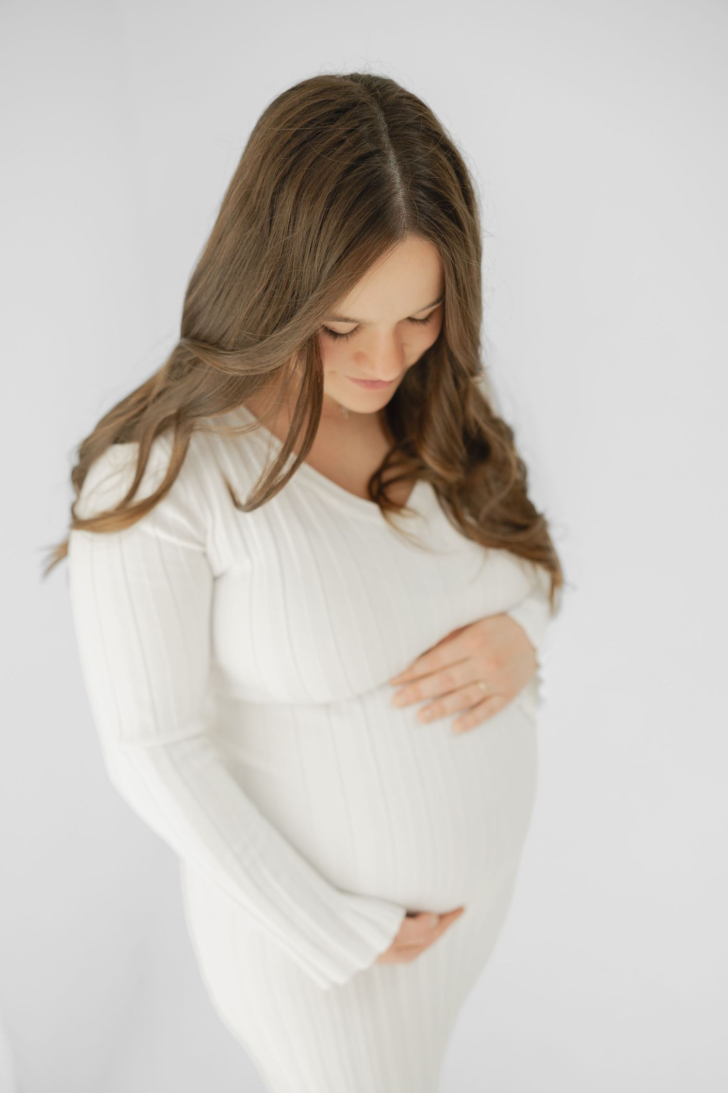 Pregnant woman in white ribbed dress cradling her baby bump, with long brown hair, against a white background.