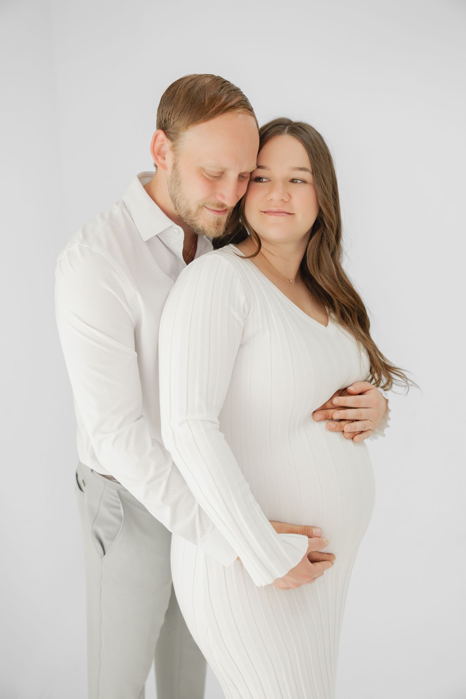 A couple in white outfits poses tenderly, the man embracing a pregnant woman from behind against a white background.
