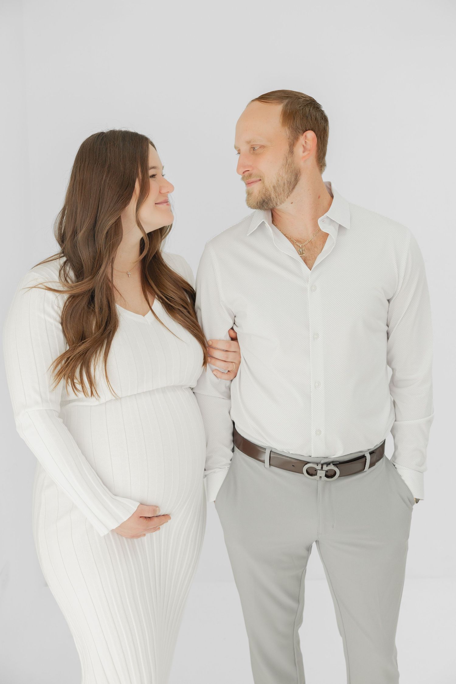 Expecting couple in white outfits smiling at each other, woman in ribbed dress cradling baby bump in studio photo.