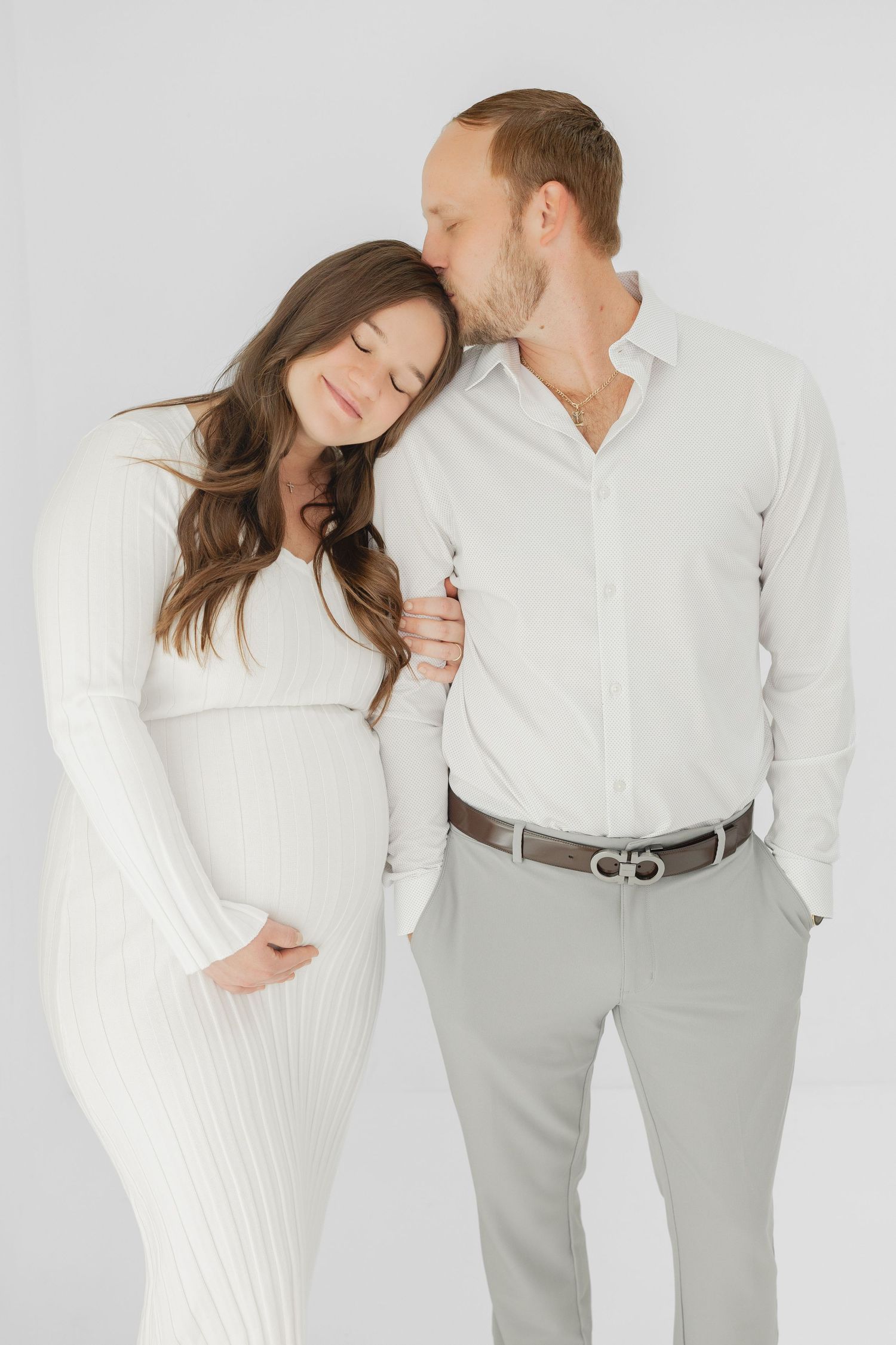 A man tenderly kisses a pregnant woman on the head as she leans against him, both dressed in white.