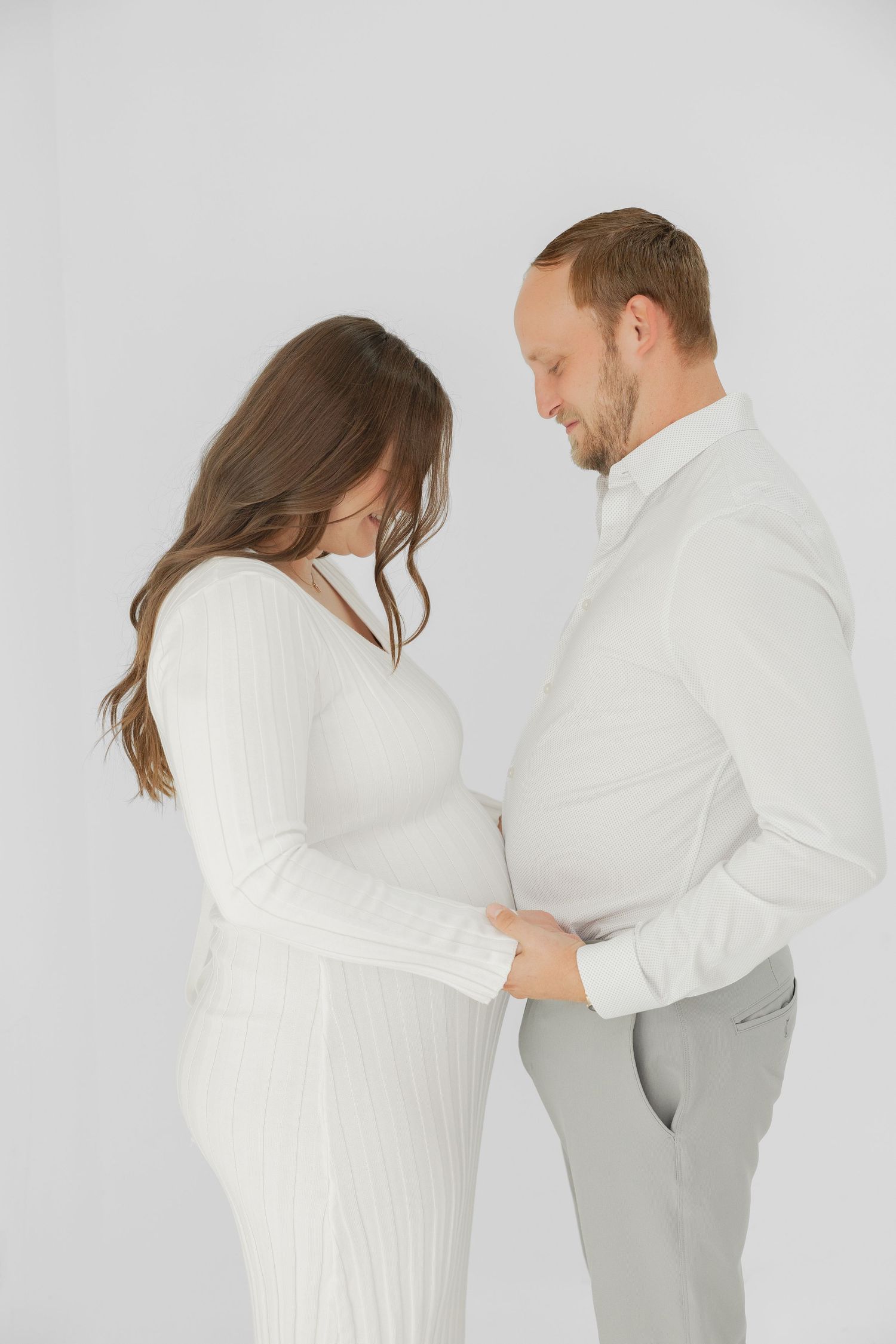 Couple in white outfits sharing a tender moment, man smiling at pregnant woman's belly in a studio setting.