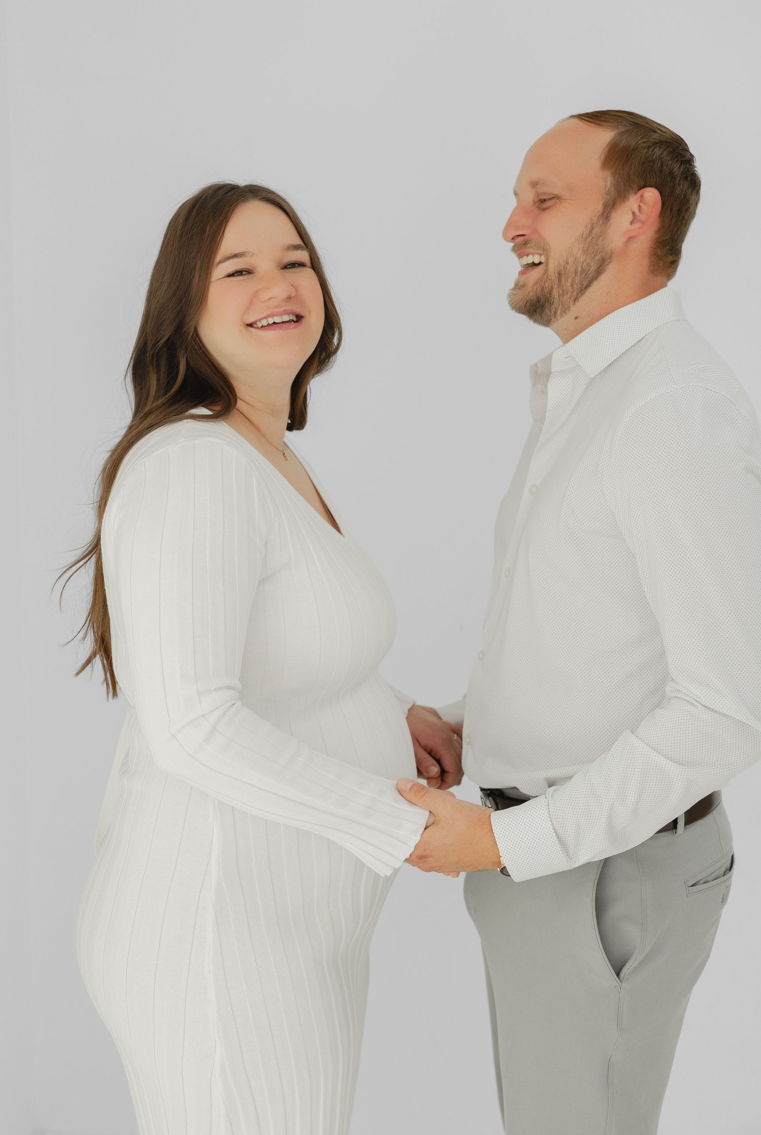 Smiling pregnant couple in white outfits holding hands against a light gray background in a maternity photo shoot.