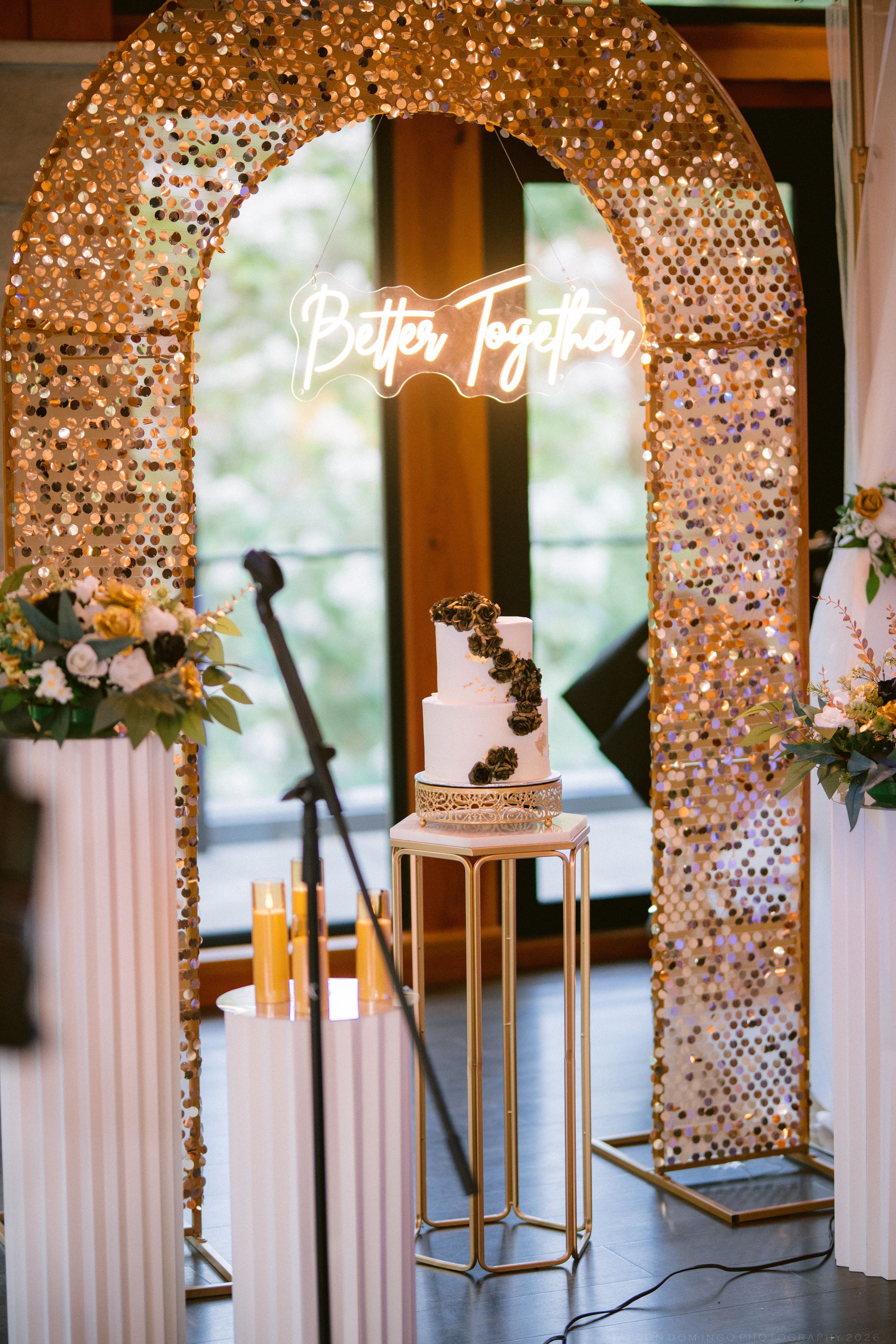 Elegant wedding cake display under a glittering gold sequin arch with a neon Better Together sign.