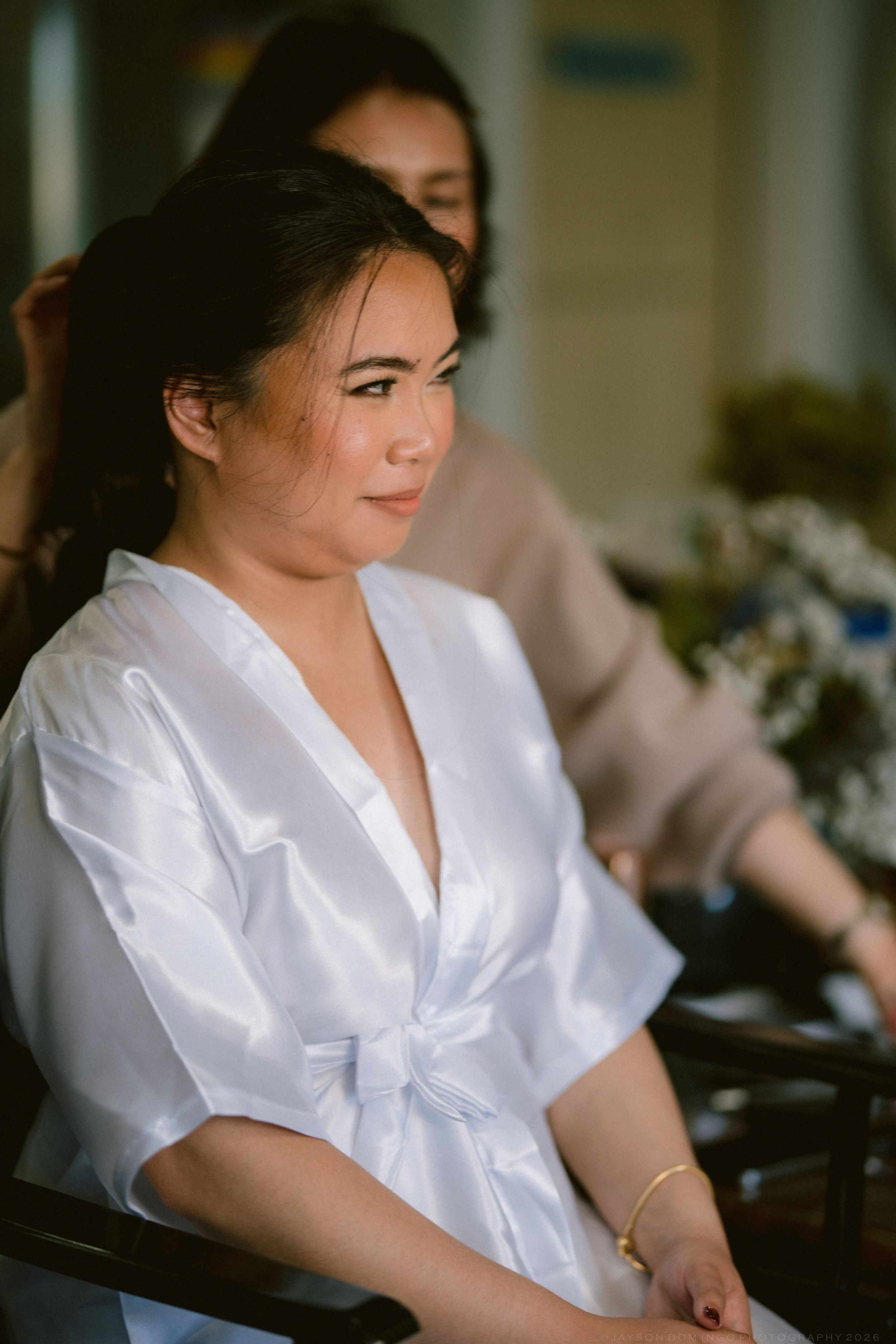 Bride in white satin robe getting hair done before wedding ceremony.