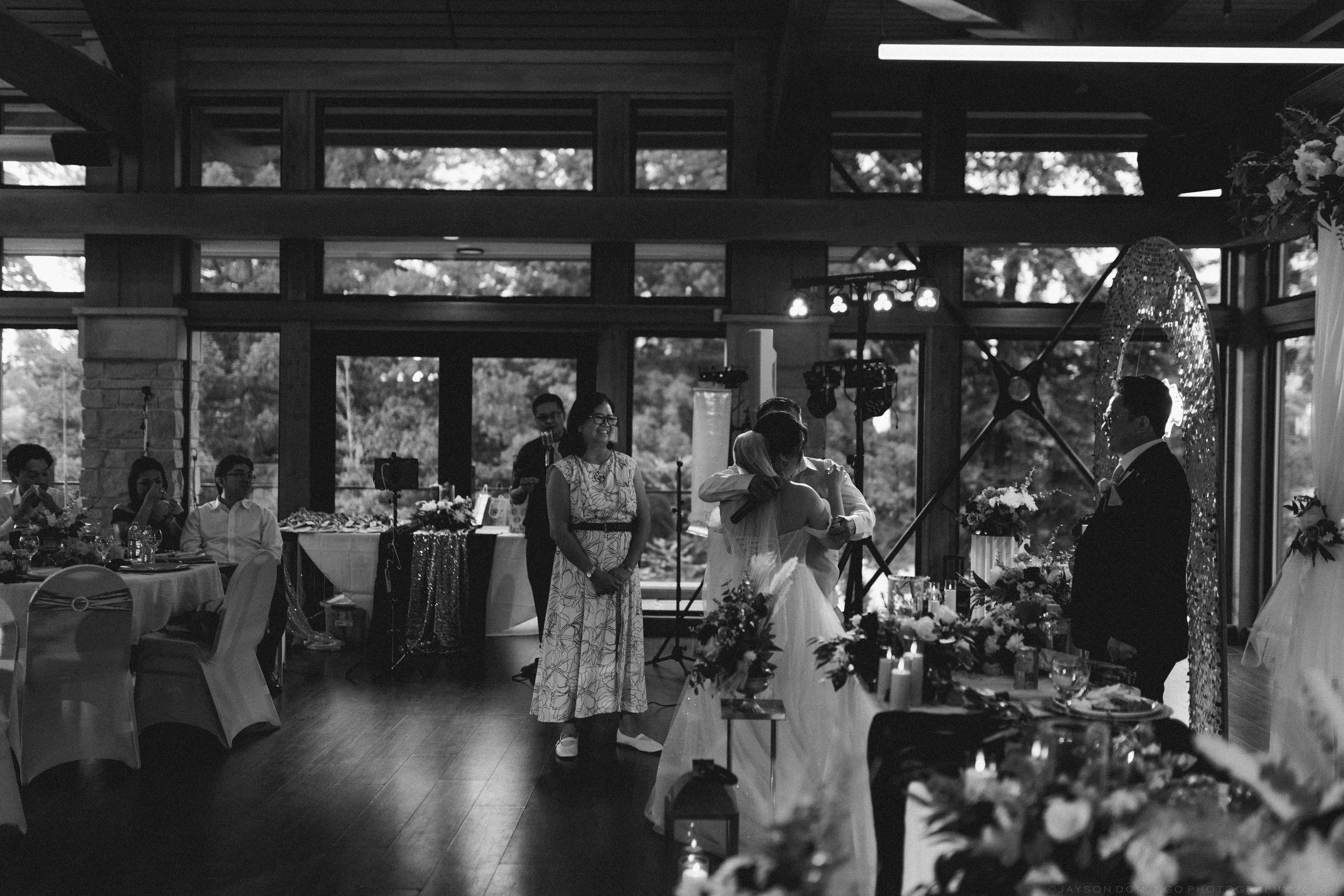 Black and white photo of wedding guests gathered in a sunlit reception hall during a speech.