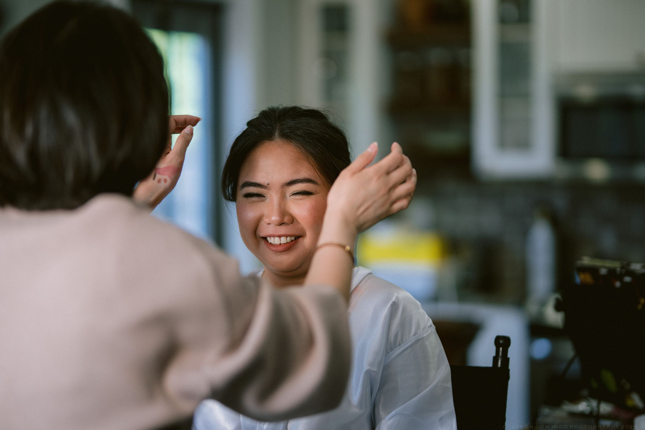 A smiling bride gets her hair styled by a makeup artist during wedding preparations.