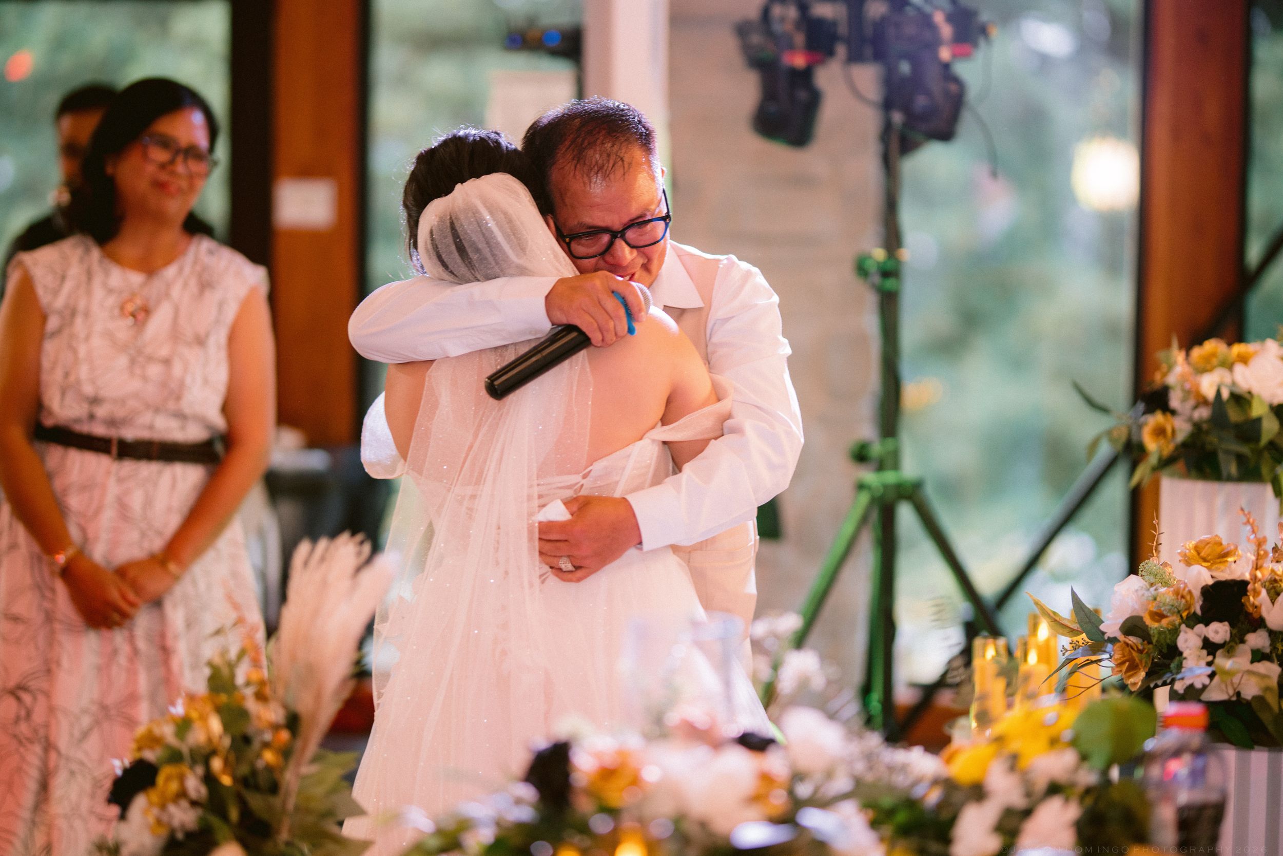 A bride in a white gown shares an emotional hug with a man holding a microphone at a wedding.