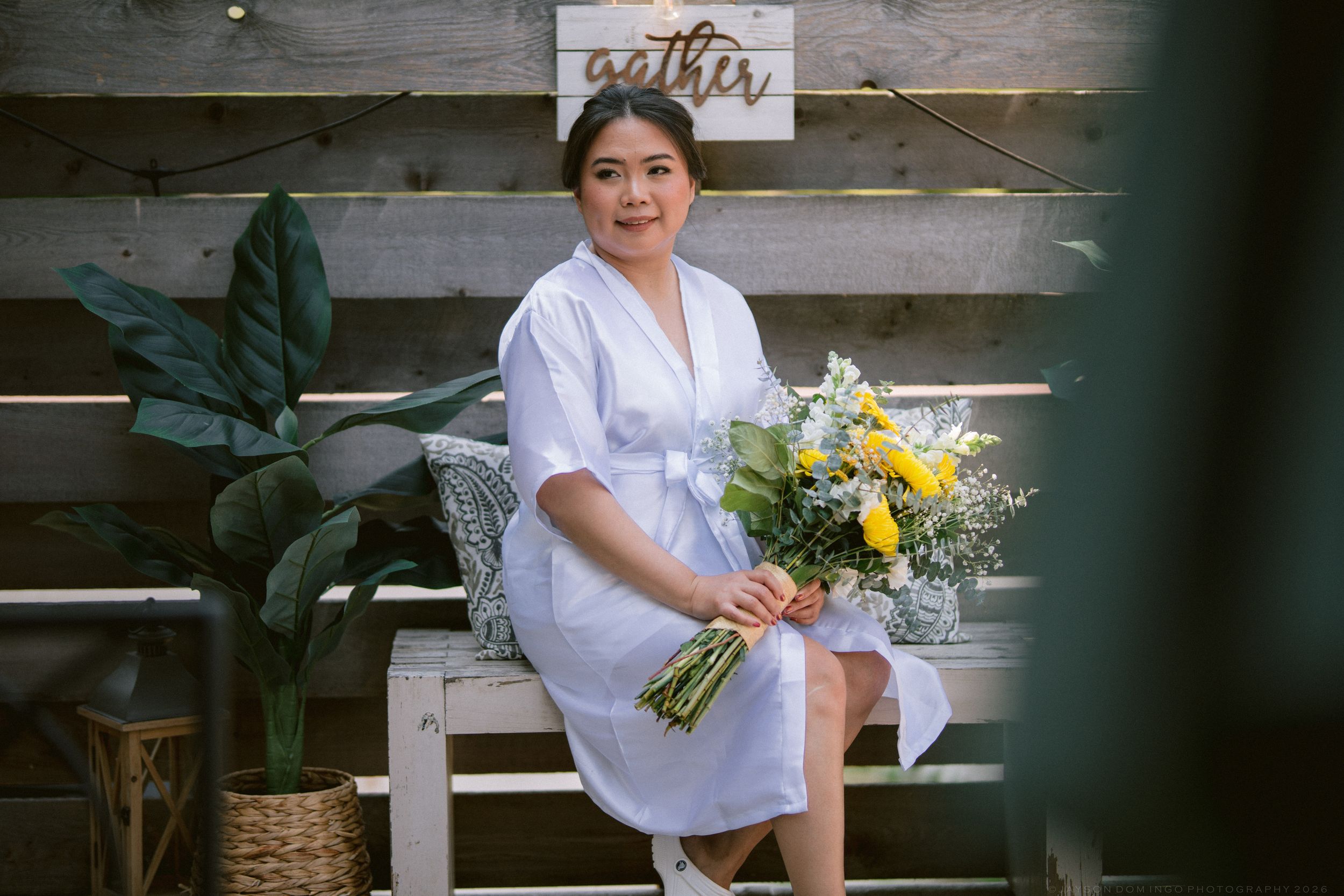 Woman in white dress sitting on bench holding yellow and white flower bouquet outdoors near wooden wall.