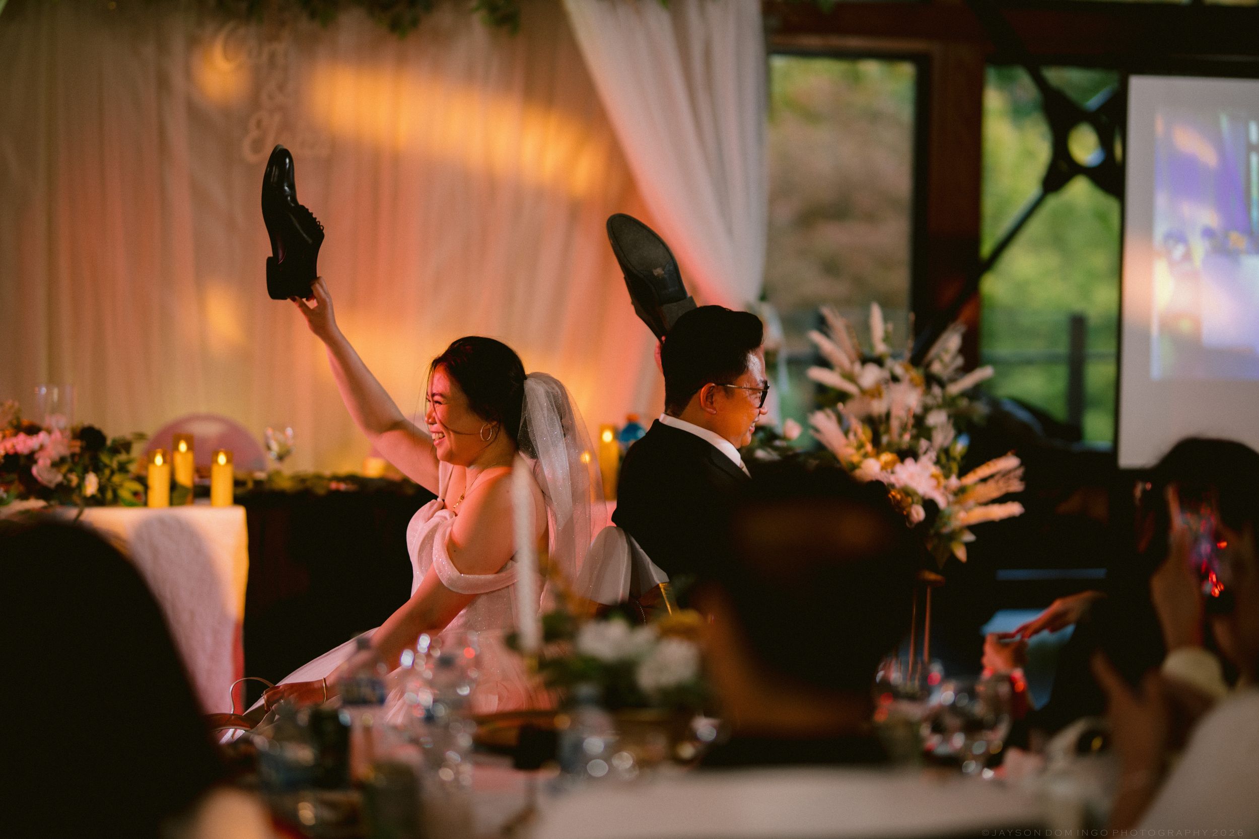 Bride raises shoes while groom sits beside her at decorated wedding reception head table.