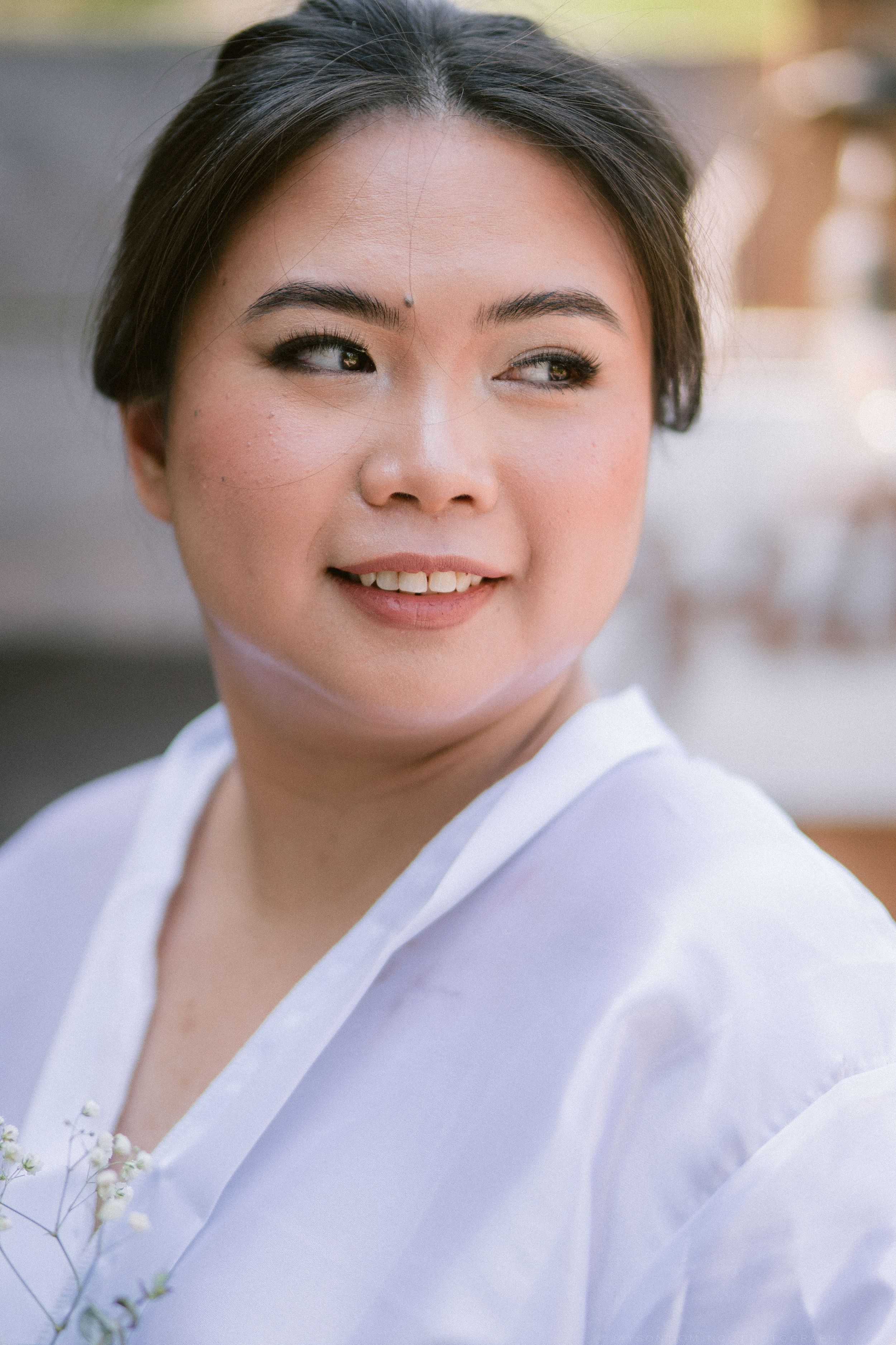 Smiling Asian woman in white outfit holding small white flowers, portrait shot with soft bokeh background.