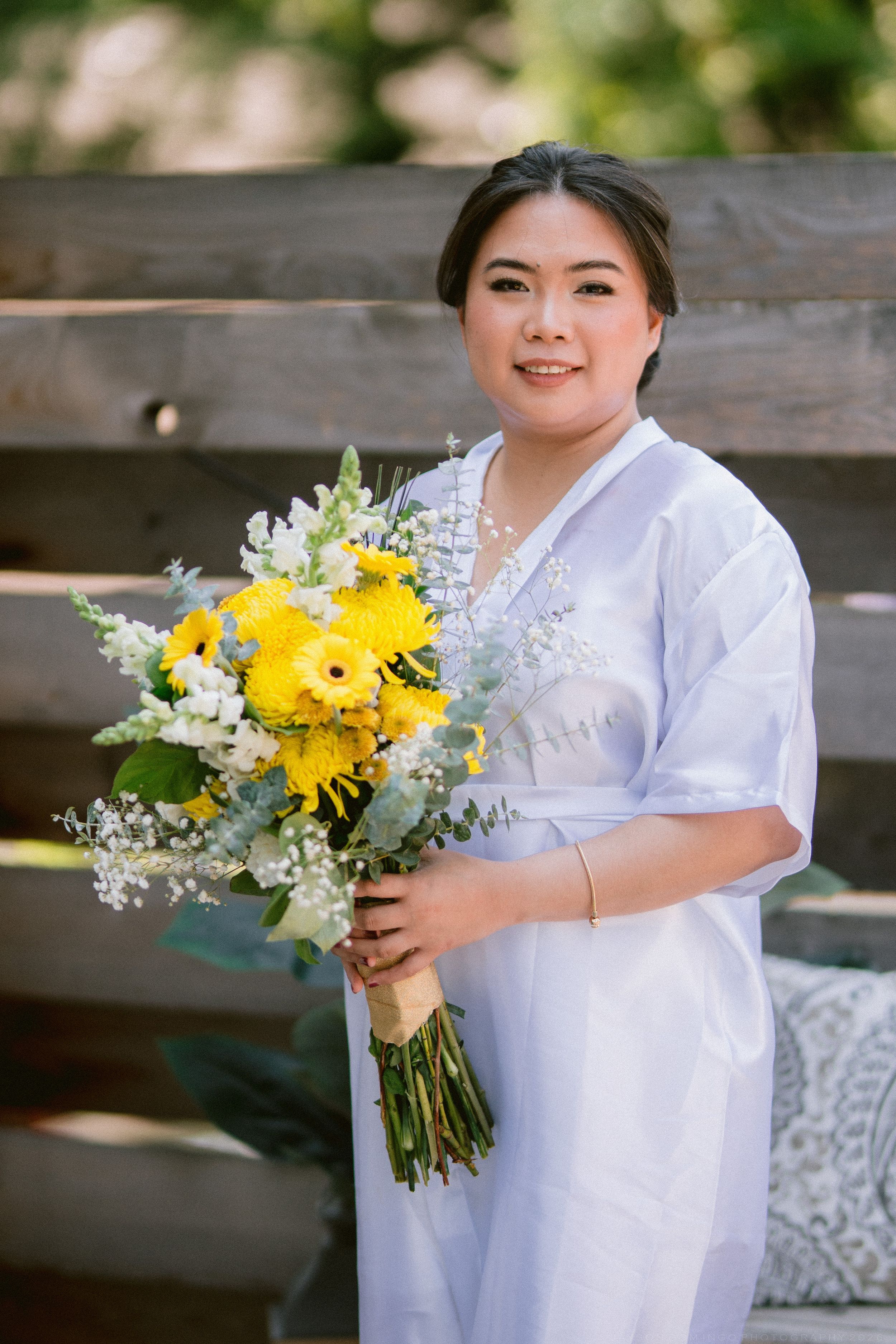Smiling bride in white dress holds vibrant yellow and white flower bouquet outdoors near wooden fence.