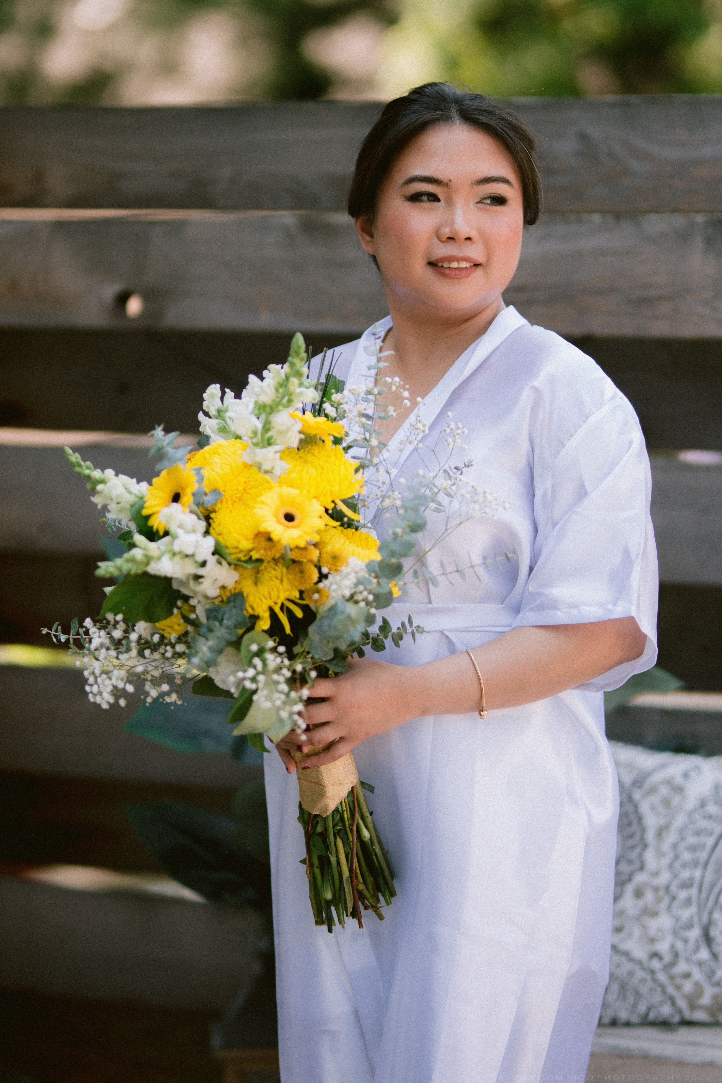 Asian bride in white dress smiles holding yellow sunflower and white floral bouquet near wooden backdrop.