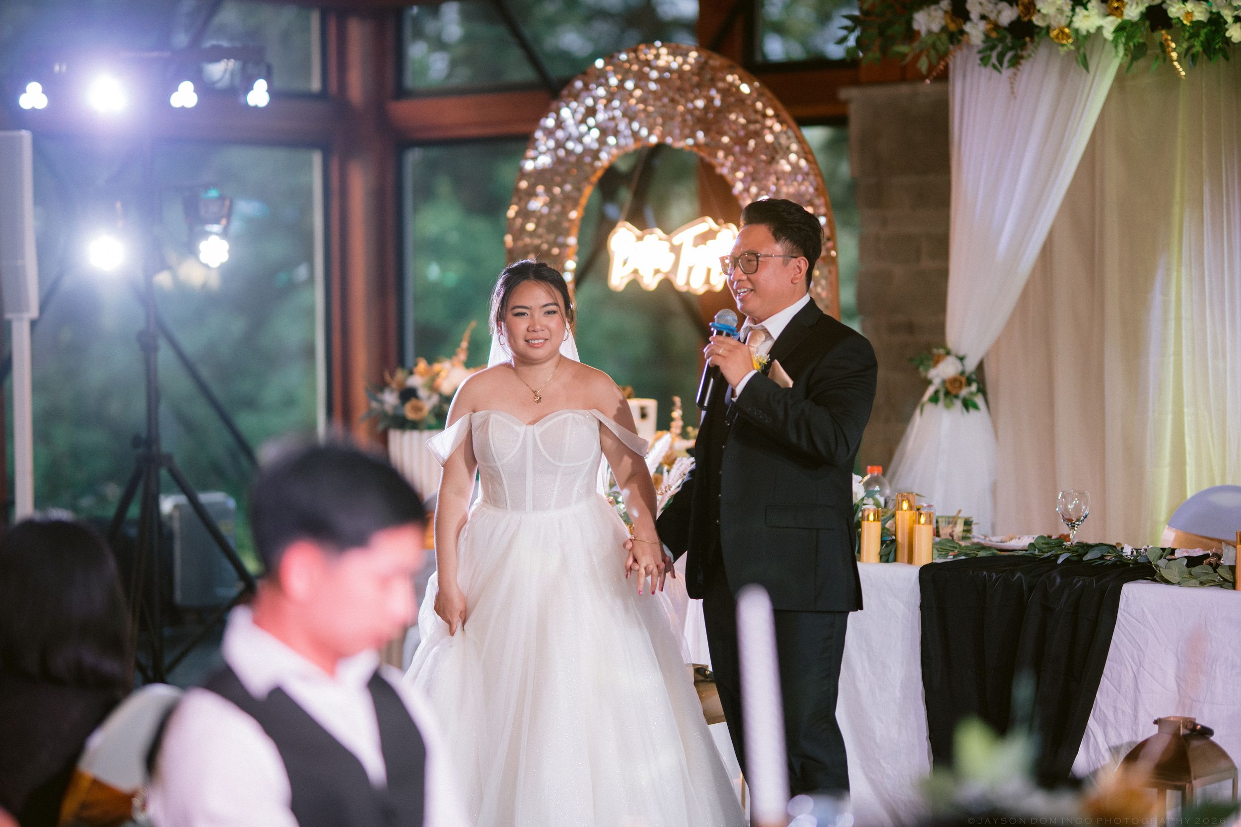Bride and groom standing at floral arch with string lights during wedding reception speech.