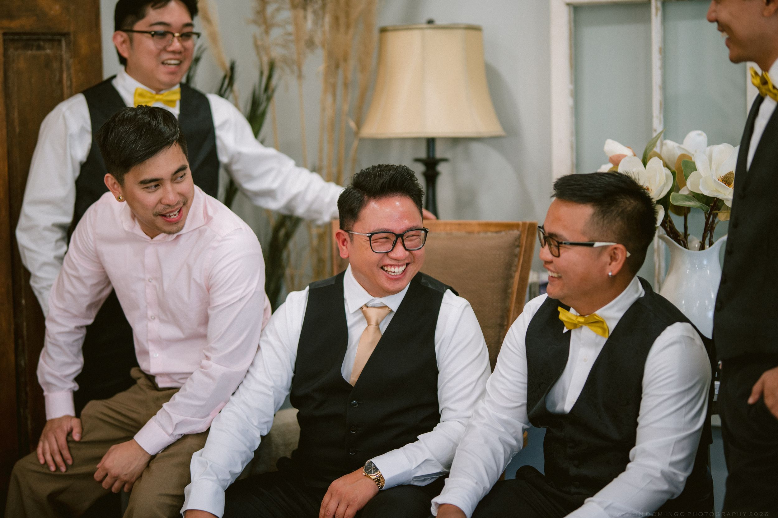 Groomsmen in white shirts, vests, and yellow bow ties laughing together during wedding preparations.