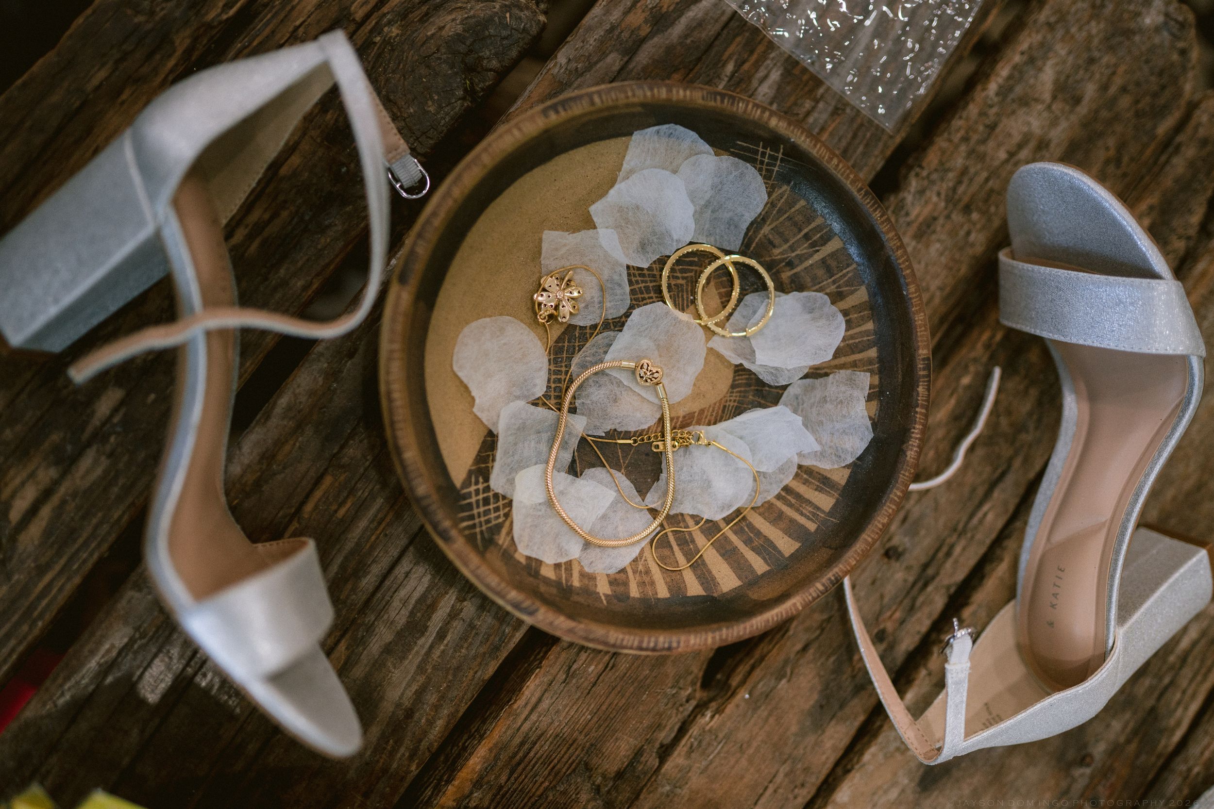 White bridal heels beside a wooden tray holding gold wedding rings and delicate floral accessories on a rustic wood surface.