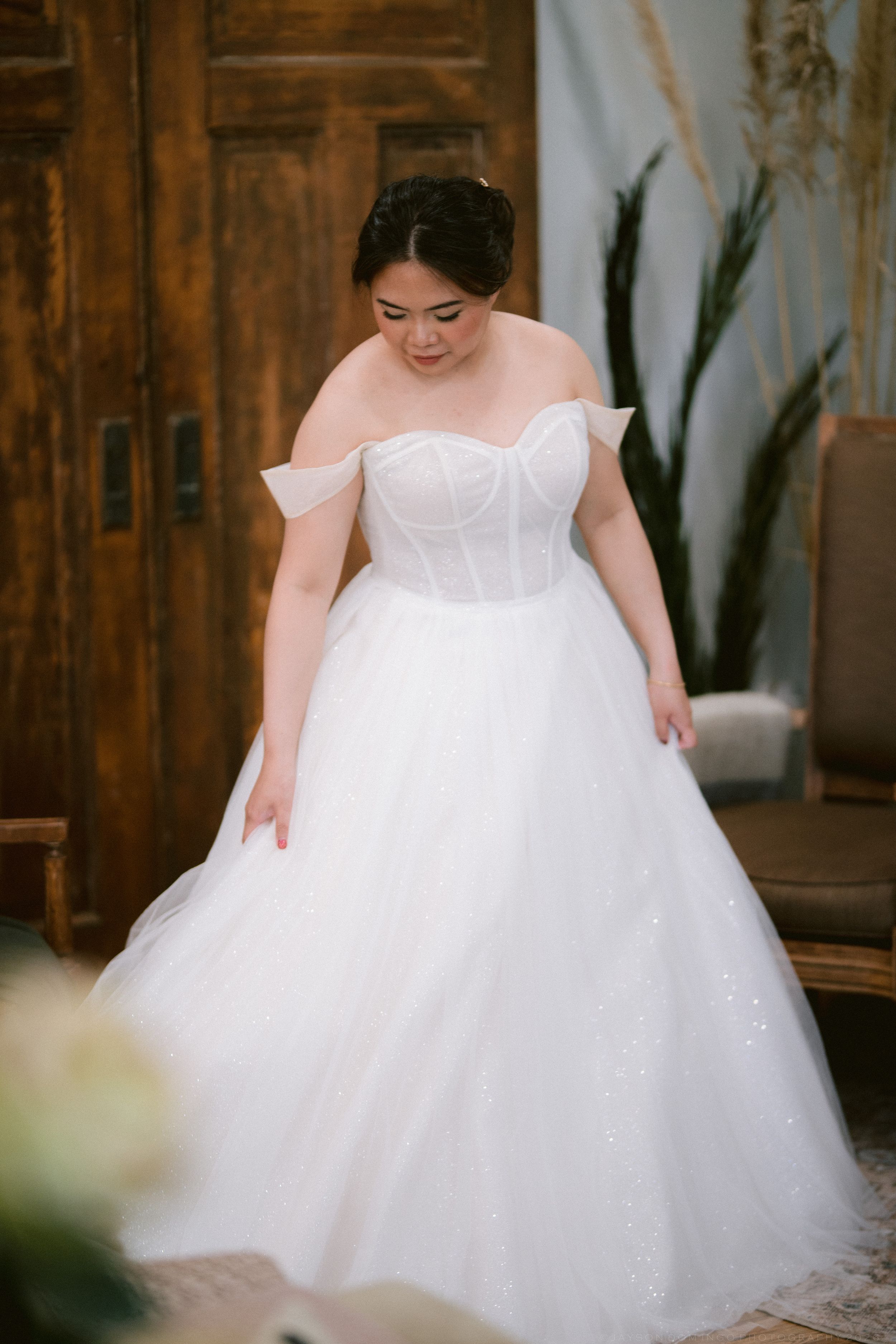 Plus-size bride in an off-shoulder white ballgown with corset bodice smiling during wedding preparation.