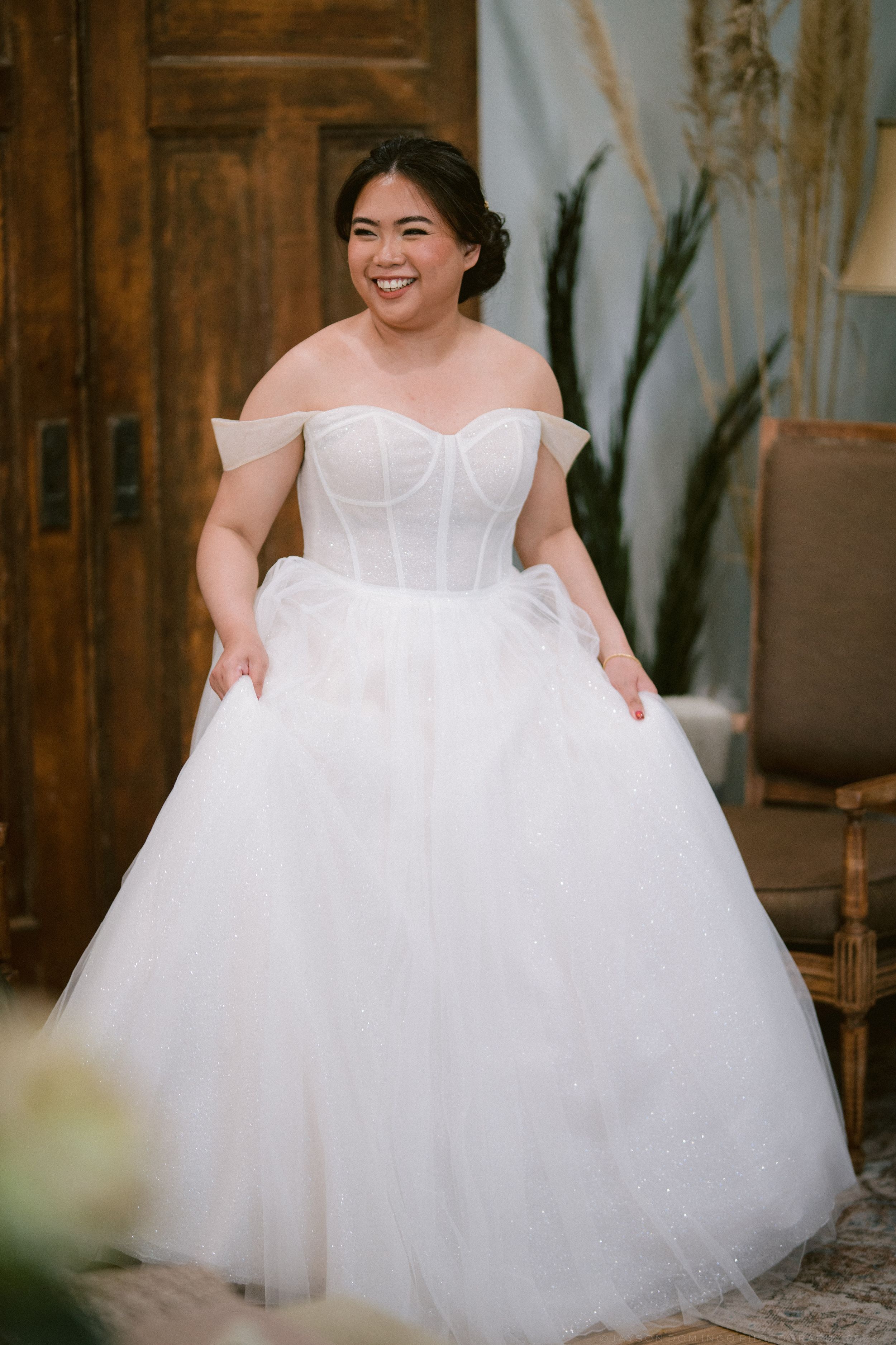 A smiling bride in an off-shoulder white corset ballgown tulle wedding dress poses indoors.