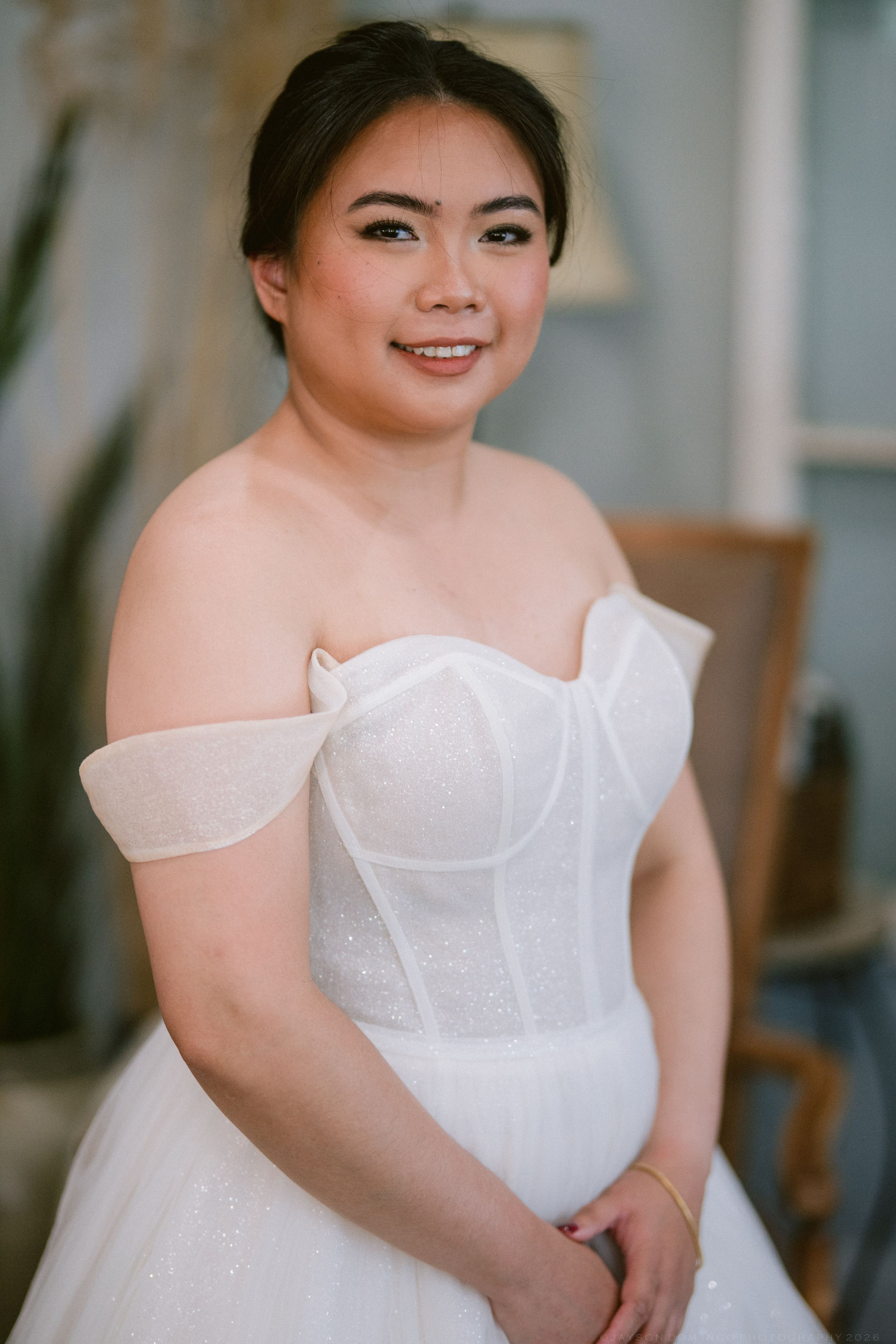 Smiling Asian bride in an off-shoulder white corset wedding gown posing elegantly indoors.