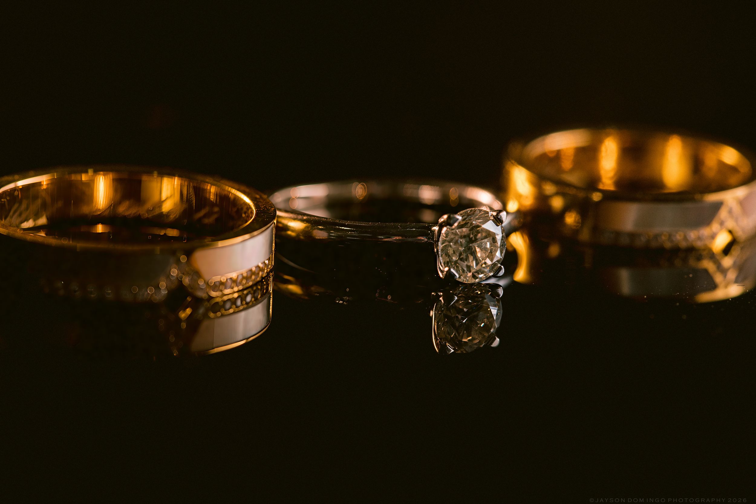 Three elegant wedding rings on a dark reflective surface, featuring two gold bands and one diamond engagement ring.