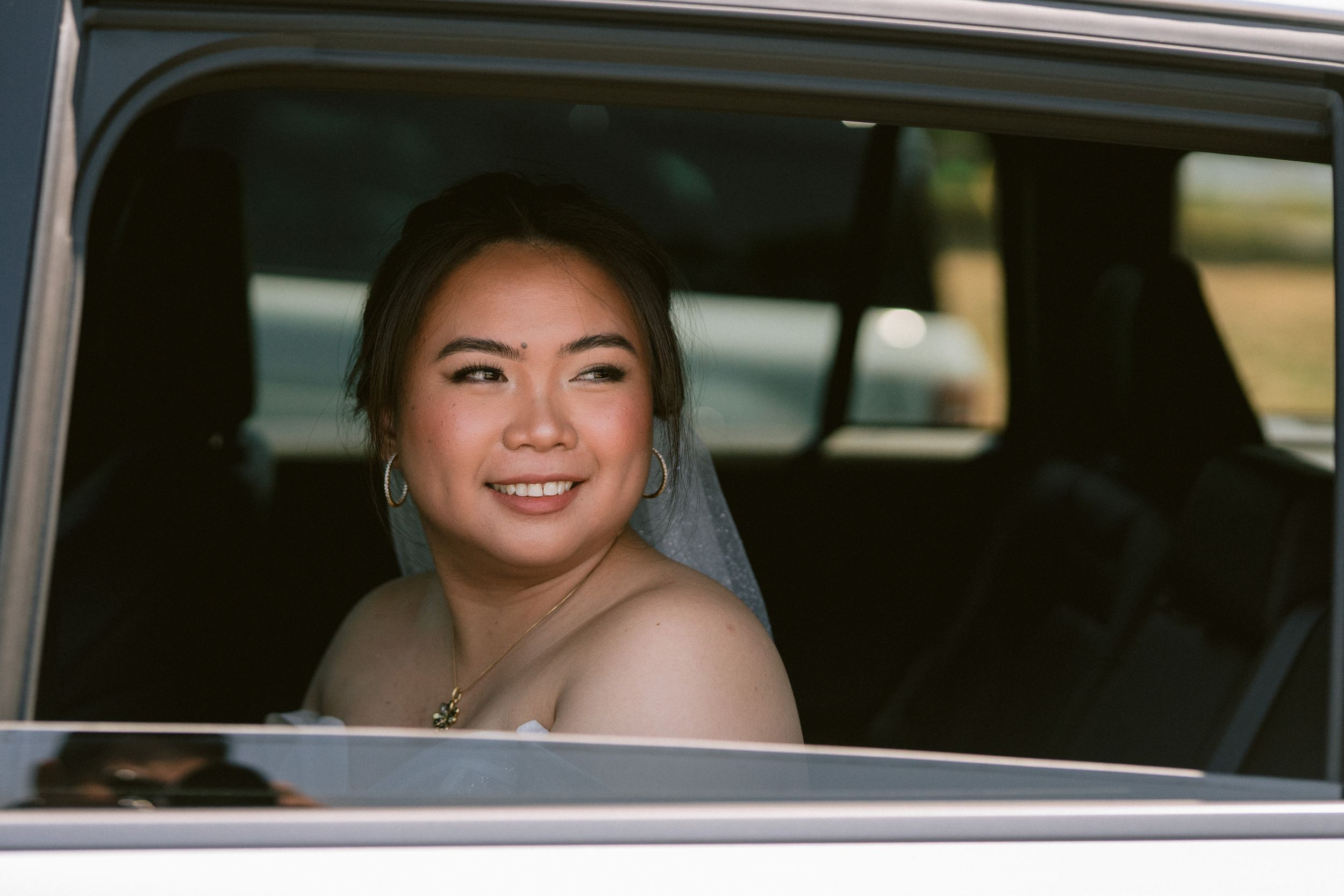 Smiling bride in a wedding veil sitting in a car, looking back over her shoulder with a joyful expression.