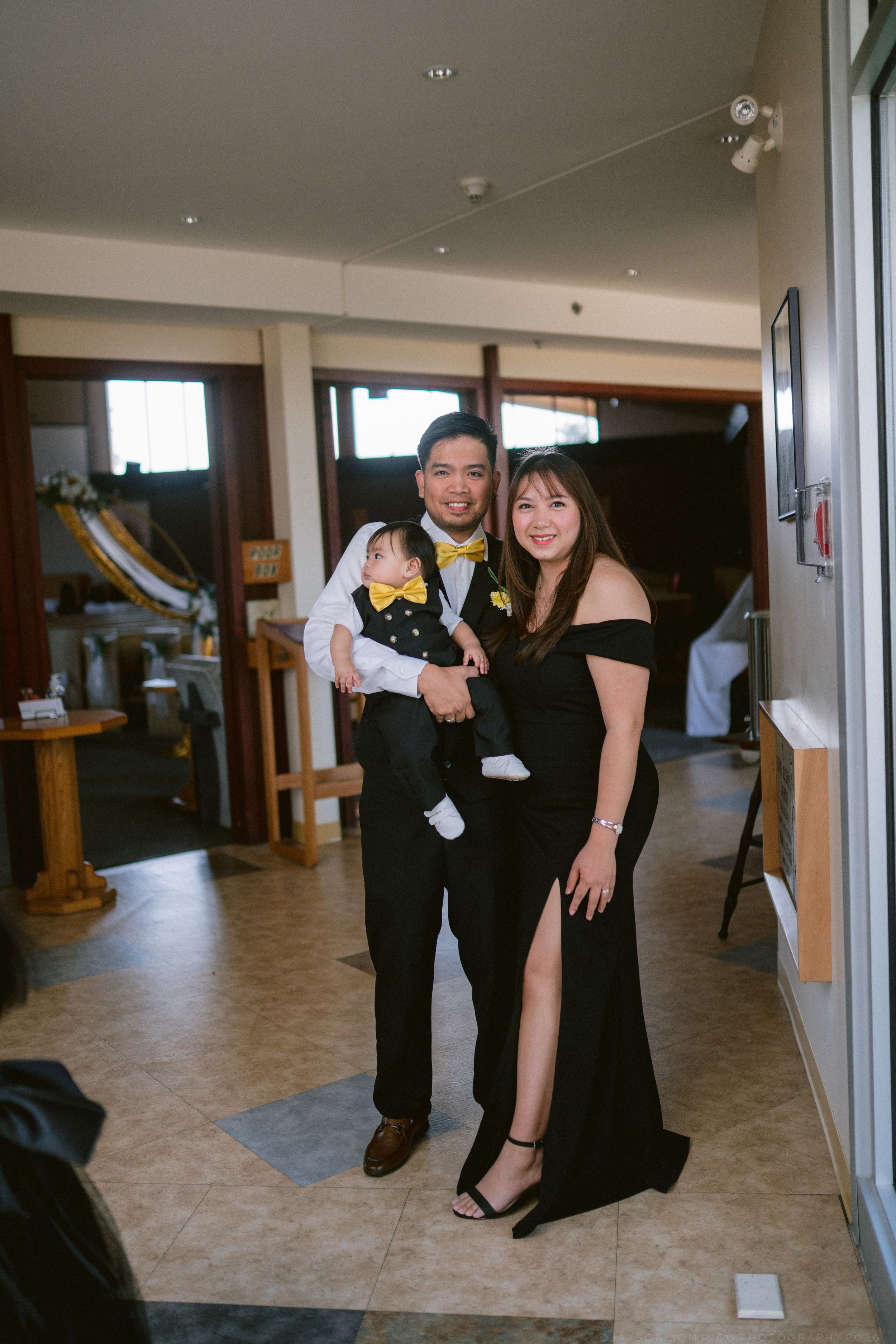 Asian family posing indoors in formal attire with matching yellow bow ties, father holding a dressed baby.