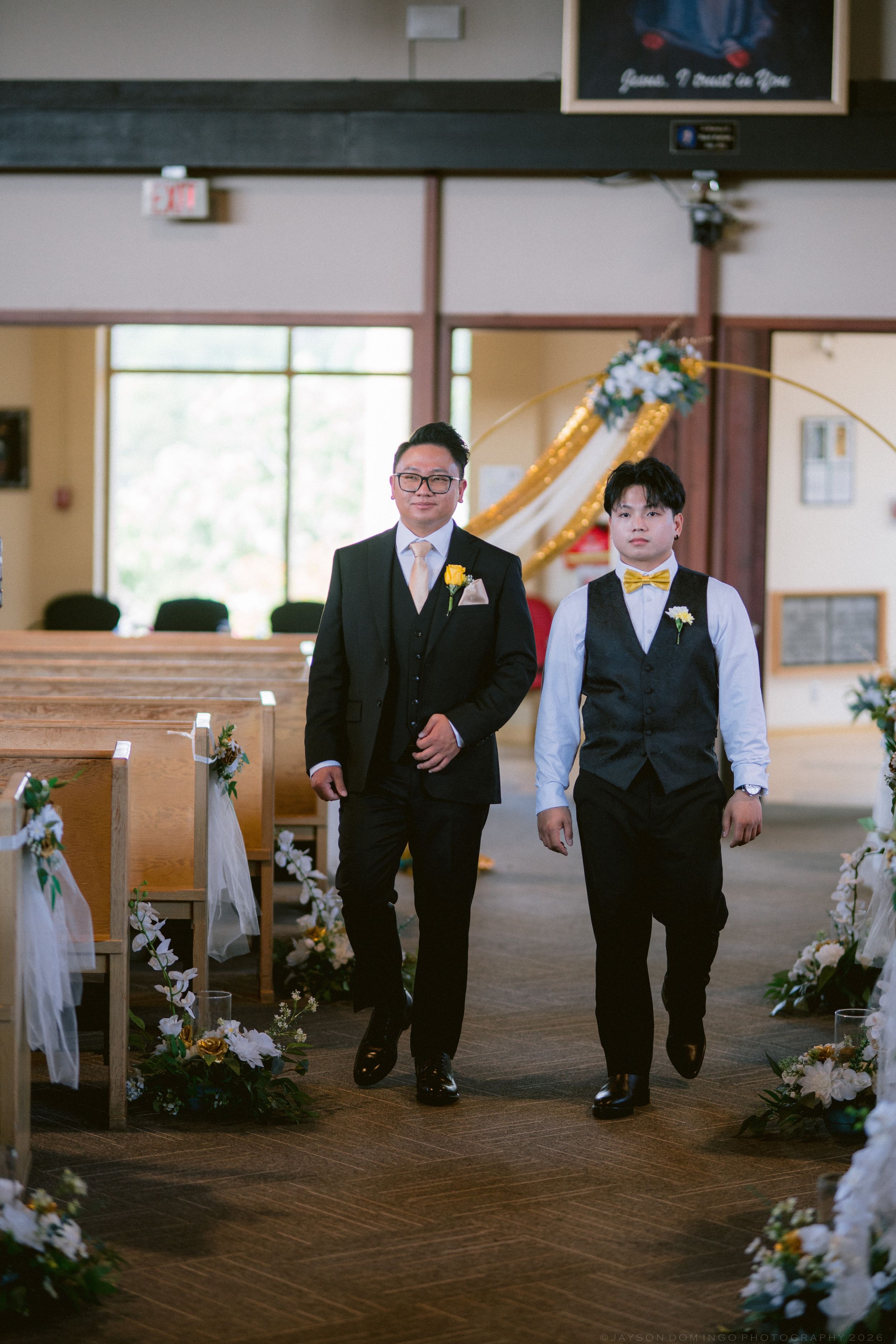 Two groomsmen in black suits with yellow boutonnieres walk down a flower-lined church aisle.