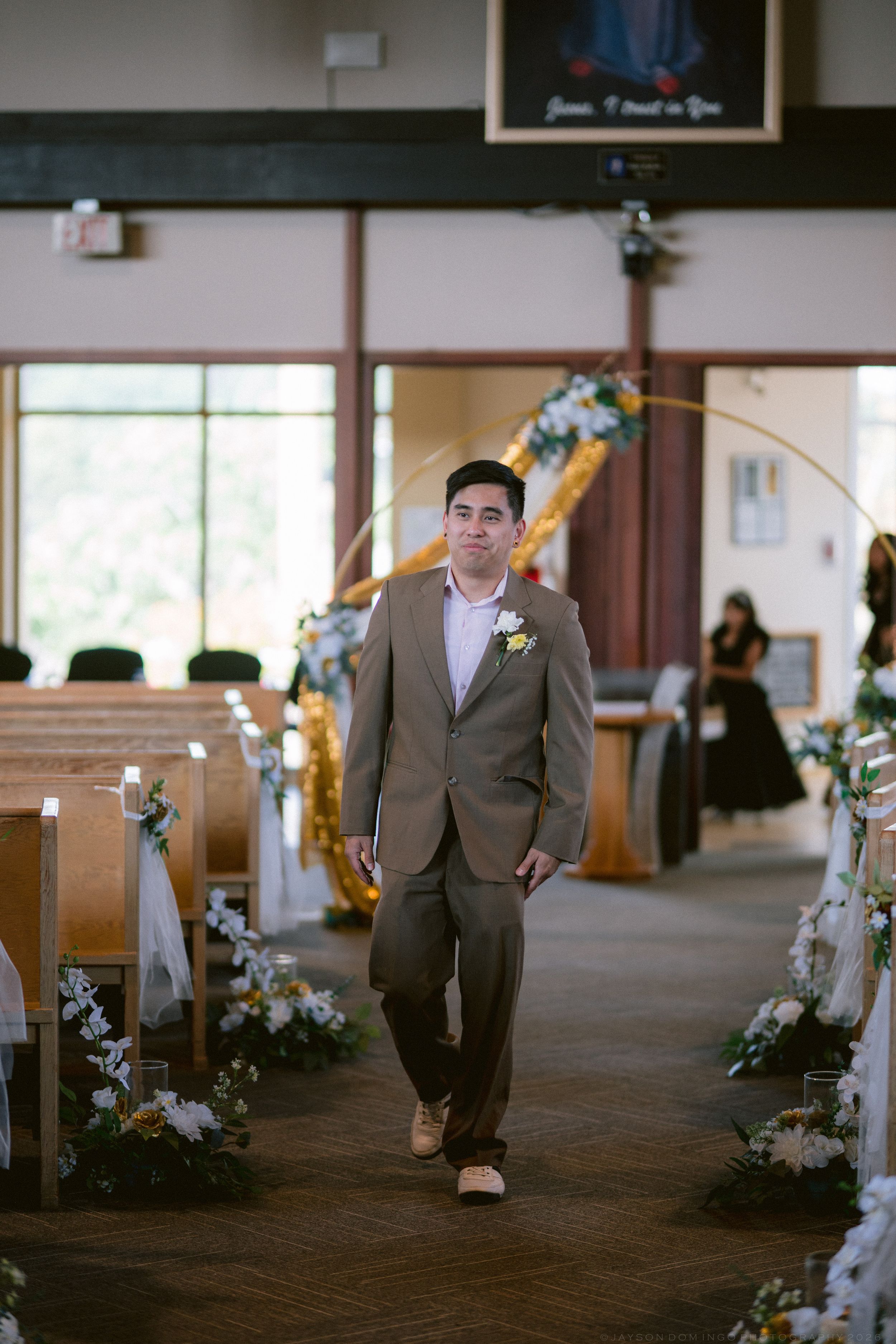 Groom in tan suit walking down decorated church aisle with floral arrangements and golden arch at altar.