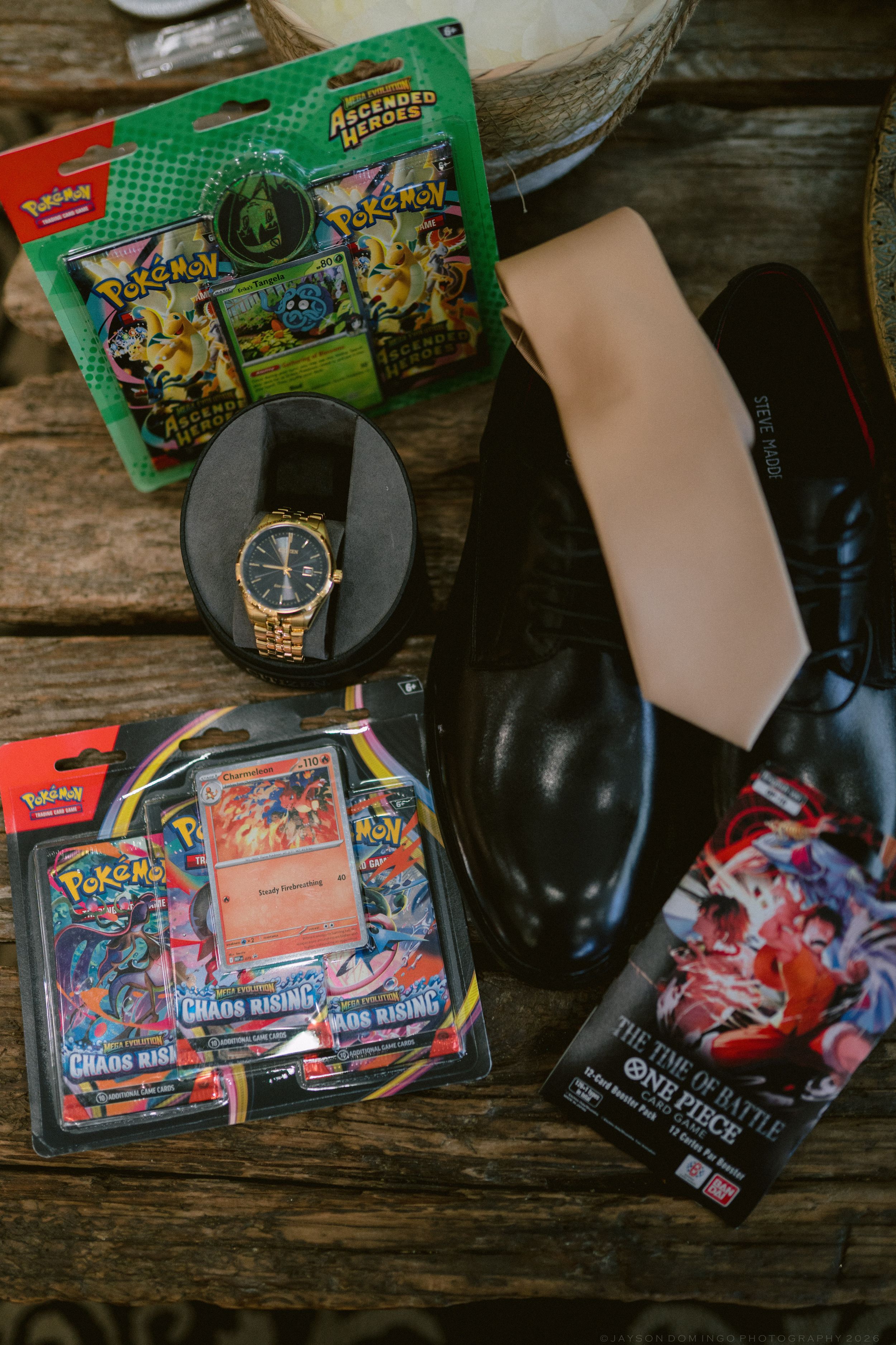 Flat lay of wedding groom gifts including Pokemon cards, a gold watch, black dress shoes, a tie, and One Piece card game.